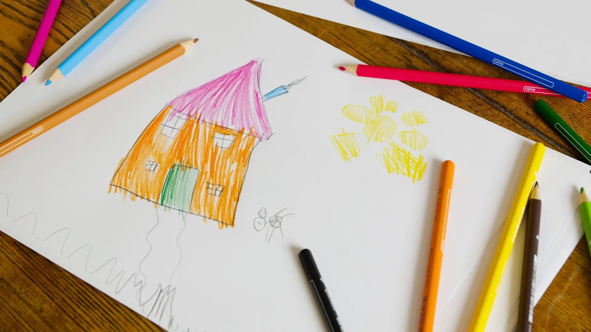 Ein Kinderzeichnung liegt mit Buntstiften auf einem Holztisch. Die Zeichnung zeigt ein Haus und eine Sonne.