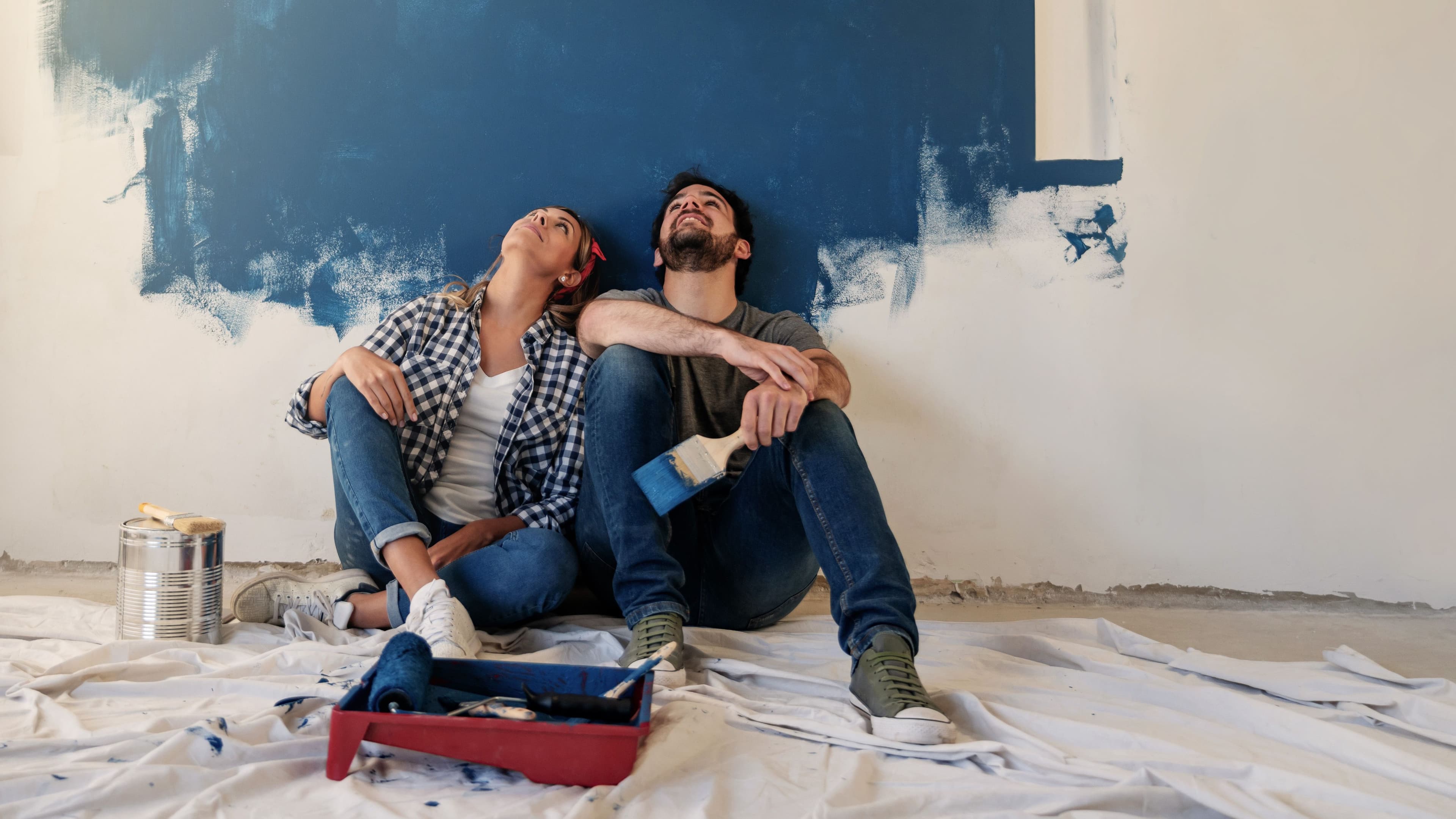 Eine Frau kniet barfuß auf einem Teppich vor einer grünen Wand, trägt eine orangefarbene Jacke und rührt Farbe in einem Eimer an. Neben ihr steht eine Leiter.