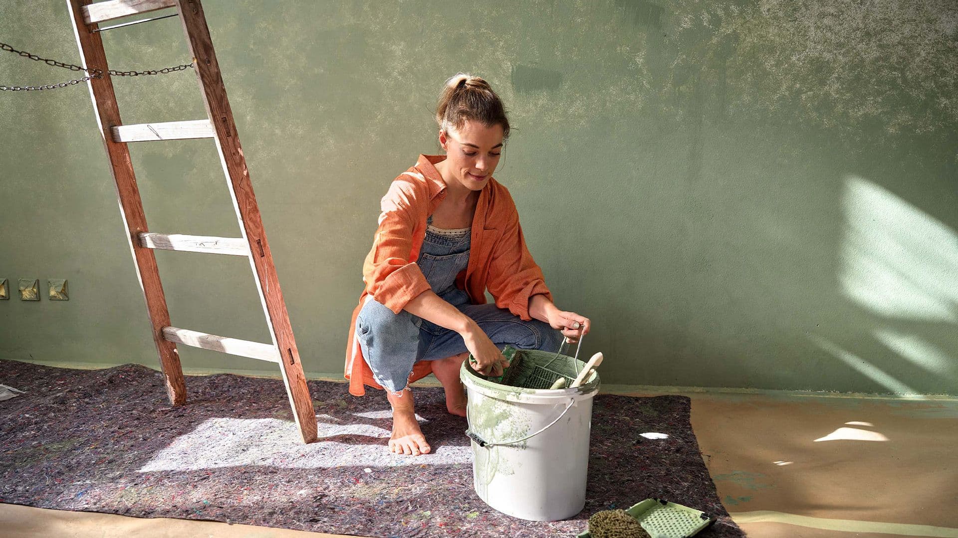 Eine Frau kniet barfuß auf einem Teppich vor einer grünen Wand, trägt eine orangefarbene Jacke und rührt Farbe in einem Eimer an. Neben ihr steht eine Leiter.
