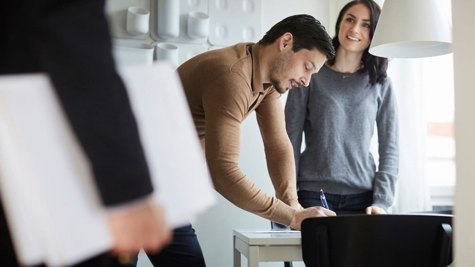 Ein Mann unterschreibt an einem Tisch ein Dokument, während eine Frau danebensteht und lächelt. Im Vordergrund hält eine weitere Person Unterlagen in der Hand. Die Szene wirkt wie ein Beratungsgespräch oder Vertragsabschluss in einem hellen Büro.