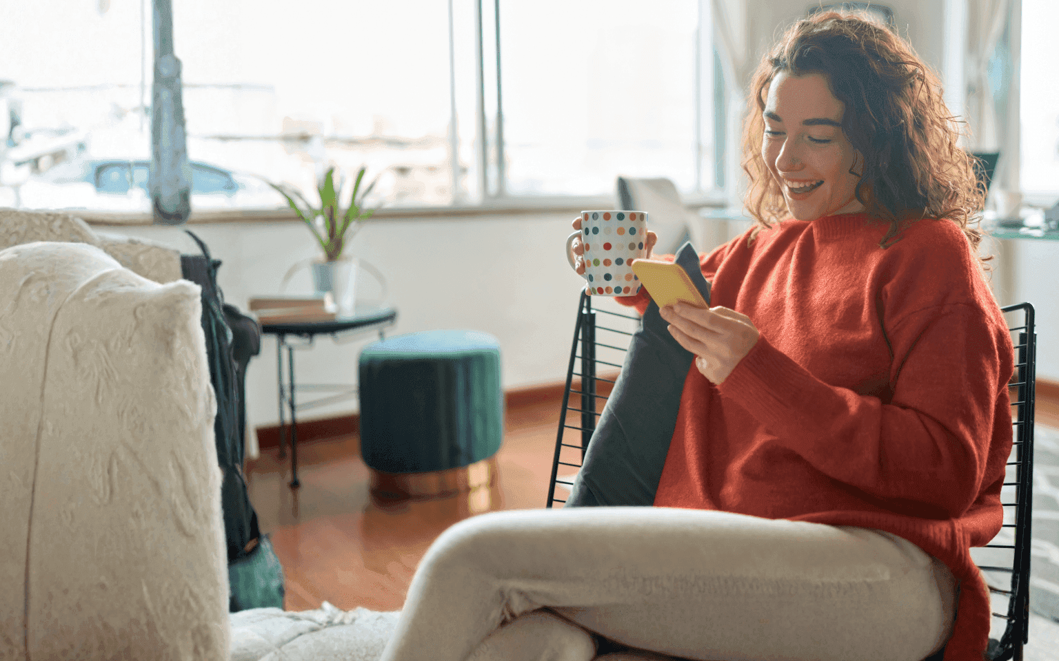 Eine junge Frau Sitzt gemütlich auf einem Stuhl in der Wonung und genießt es Kaffee zu trinken und ihr Smartphone zu nutzen.