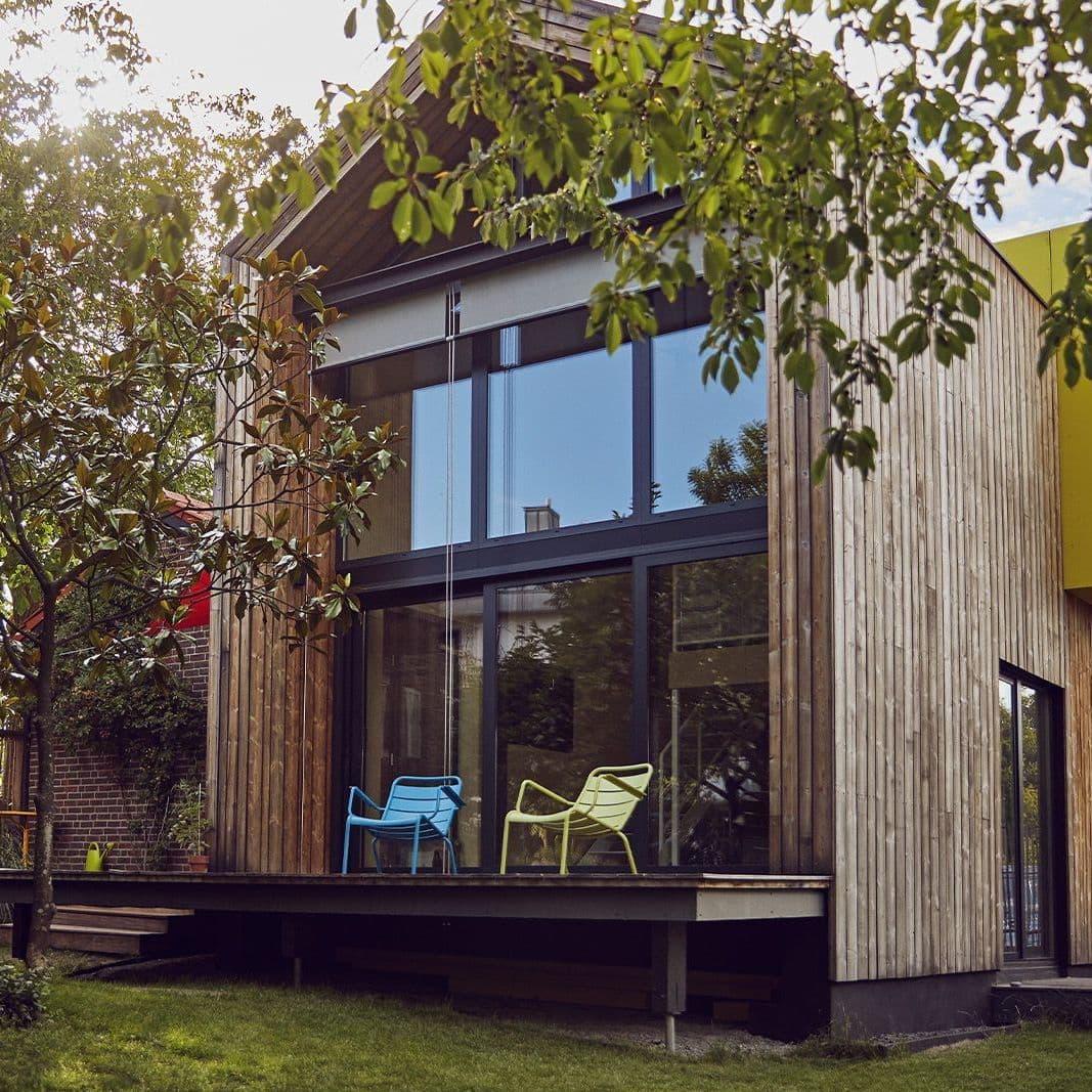 Verglaste Hausfront eines Holzhauses. Davor stehen ein blauer und ein gelber Stuhl auf einer Veranda.