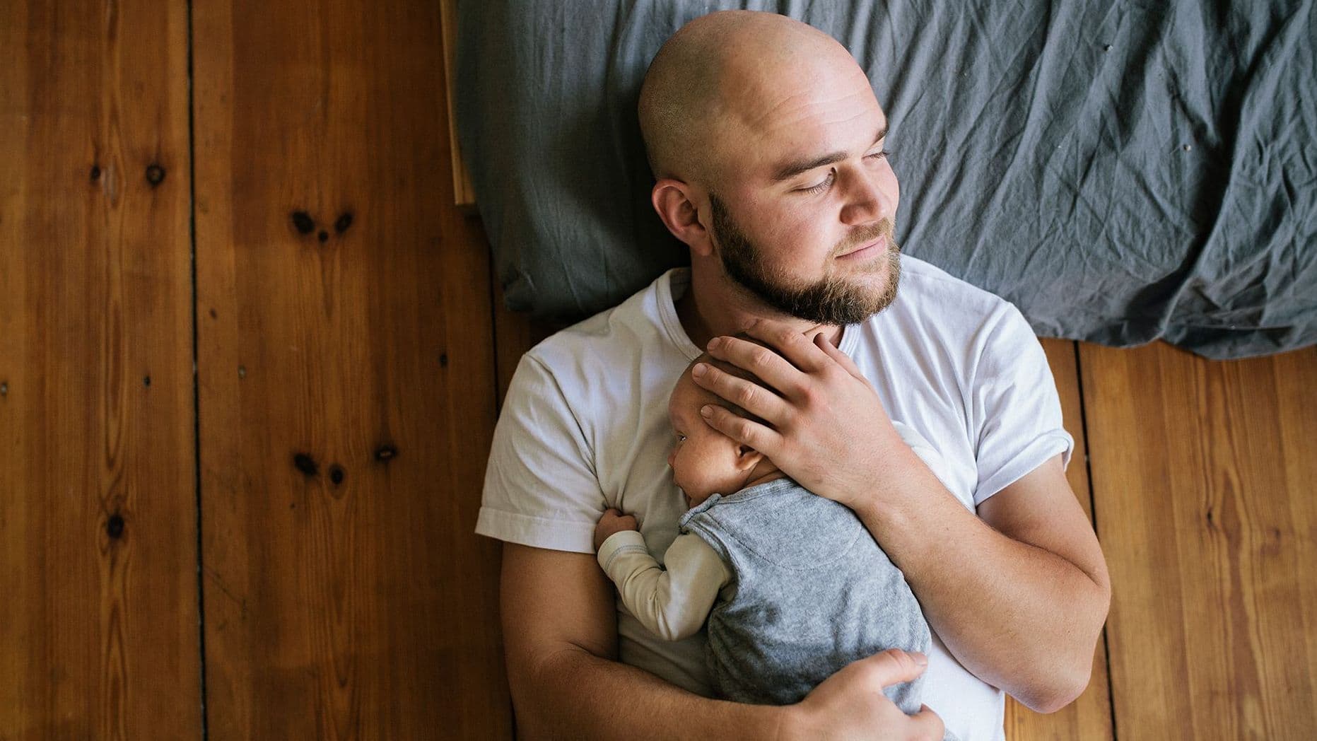 Ein Mann liegt auf einem Holzboden neben einem Bett und hält ein Baby in den Armen, während er nachdenklich zur Seite blickt.
