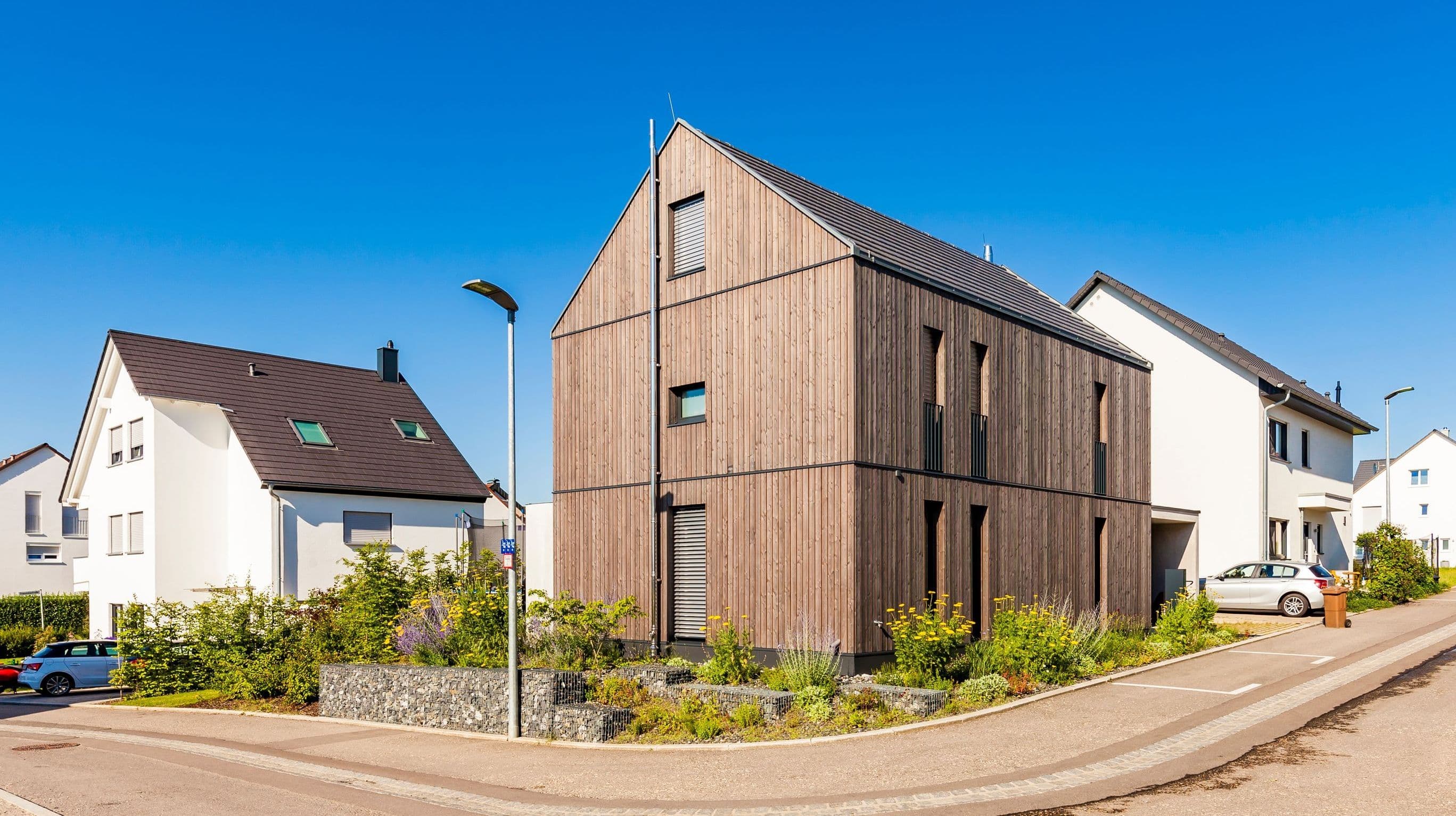 Modernes Wohnhaus mit Holzfassade an einer Straßenecke in einem Neubaugebiet bei blauem Himmel.