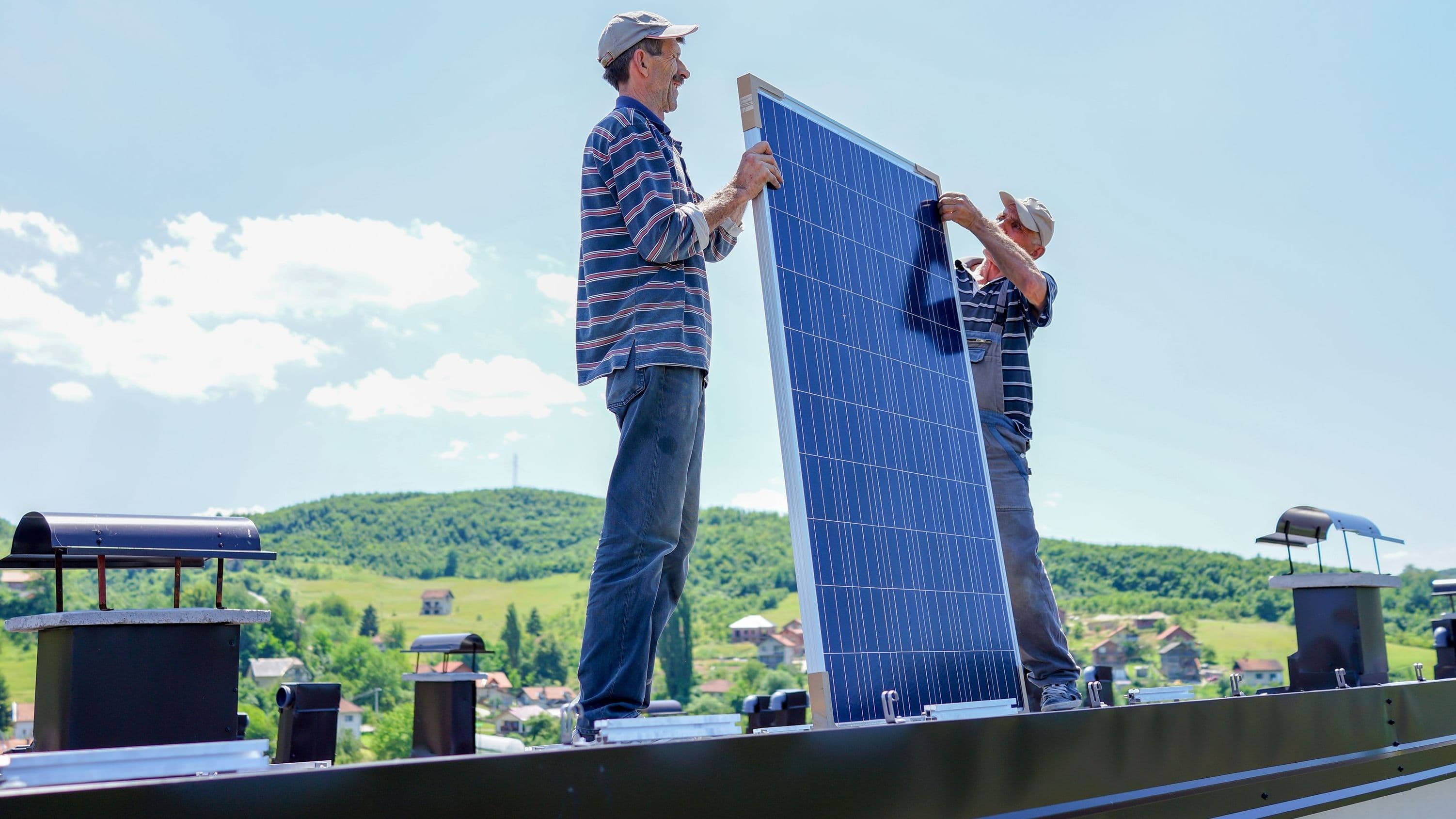 Zwei Handwerker installieren auf einem Hausdach ein Solarmodul. Sie tragen Kappen und arbeiten gemeinsam, um das Paneel auszurichten. Im Hintergrund sind grüne Hügel, Bäume und einige Häuser zu sehen.