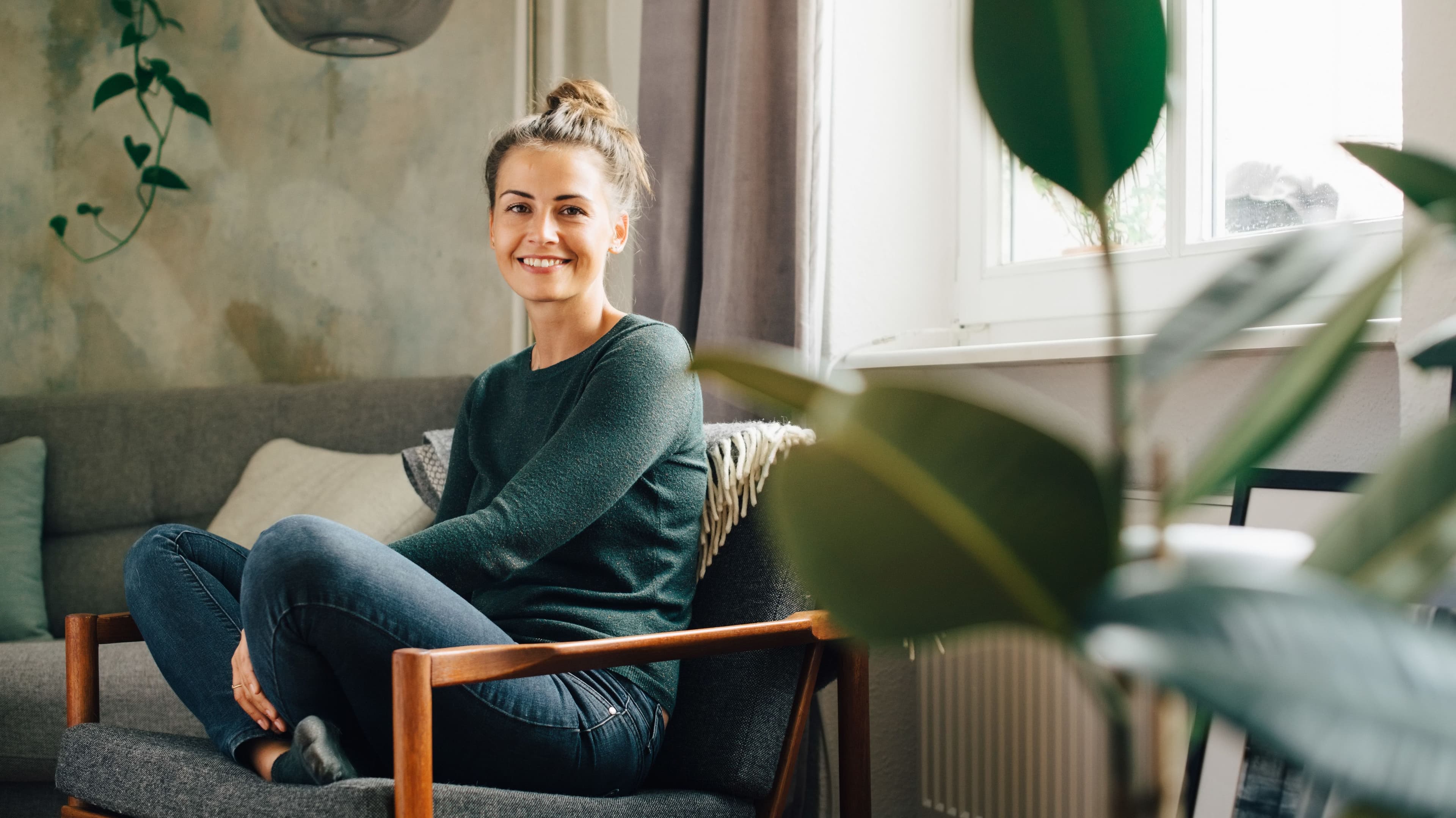Eine lächelnde Frau sitzt in einem gemütlichen Wohnzimmer auf einem grauen Sessel mit Holzrahmen. Im Vordergrund sind unscharfe Blätter einer Zimmerpflanze, im Hintergrund ein Sofa, eine Wandlampe und helle Fenster. 