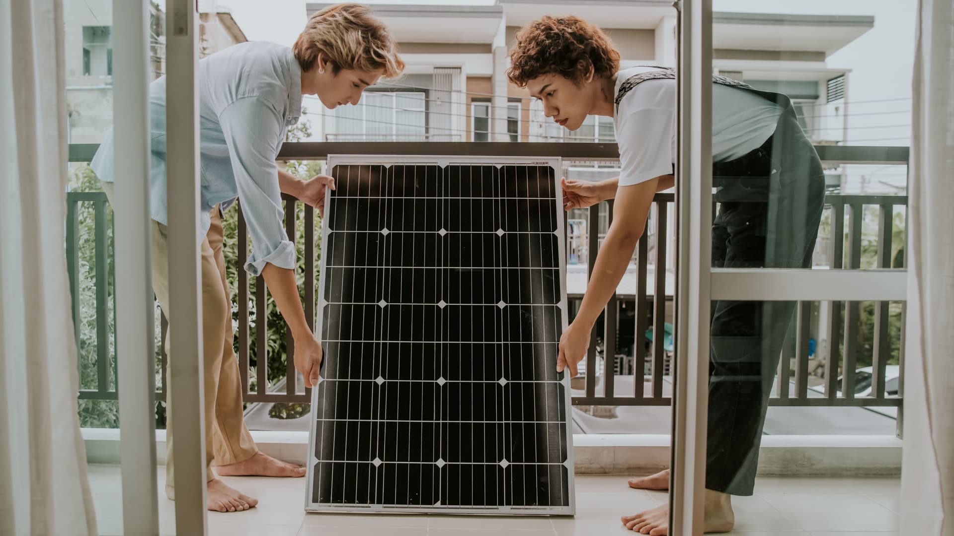 Zwei Personen tragen ein Solarpanel auf einen Balkon. Im Hintergrund sind moderne Wohnhäuser sichtbar. Die Szene spielt in einem hellen Innenraum mit offener Balkontür.