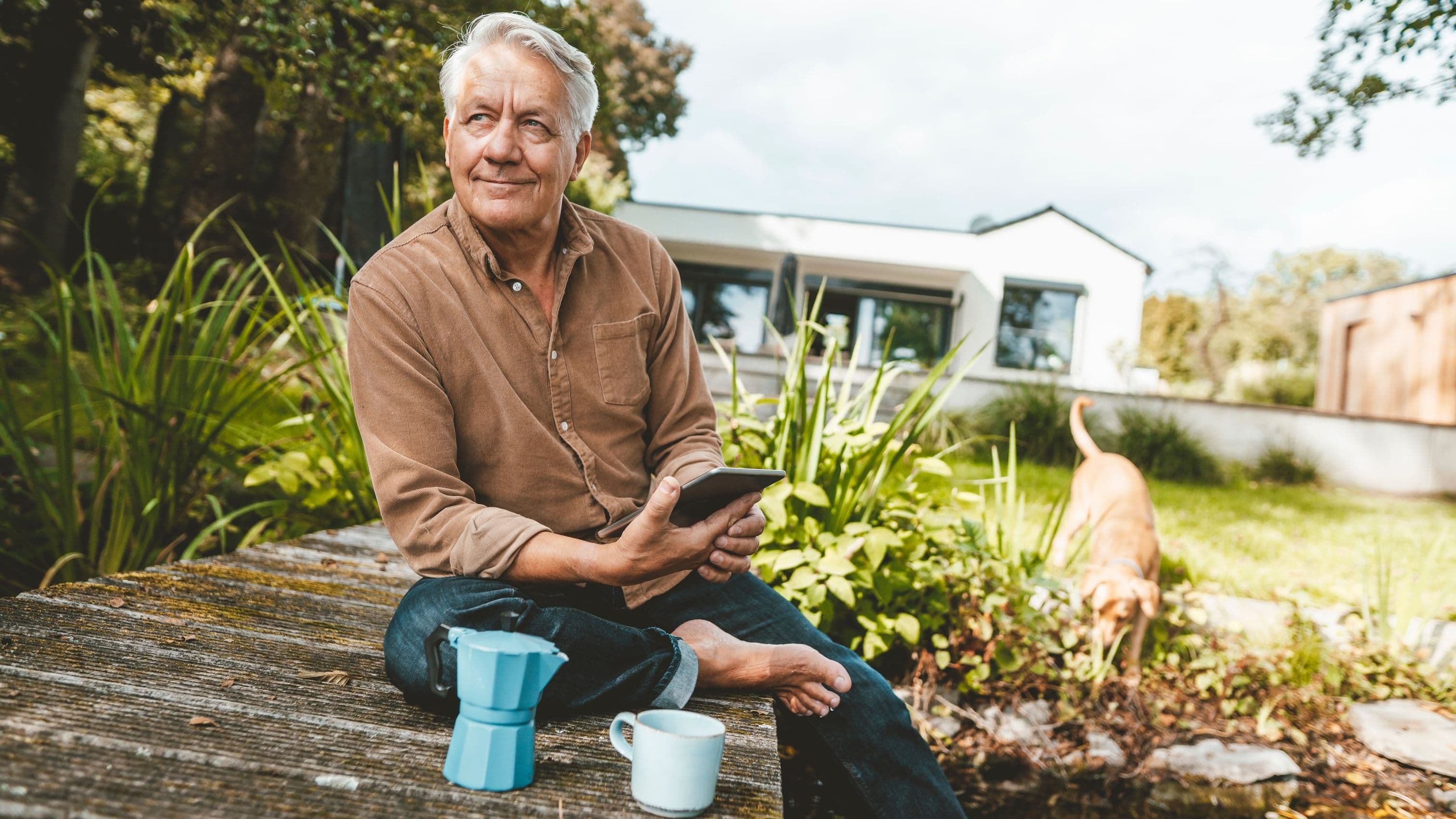 Ein älterer Mann sitzt entspannt barfuß auf einer Holzterrasse vor einem modernen Haus. In der Hand hält er ein Tablet, neben ihm stehen eine Kaffeekanne und eine Tasse. Im Hintergrund spielt ein Hund im Garten. Die Szene wirkt ruhig und naturverbunden.