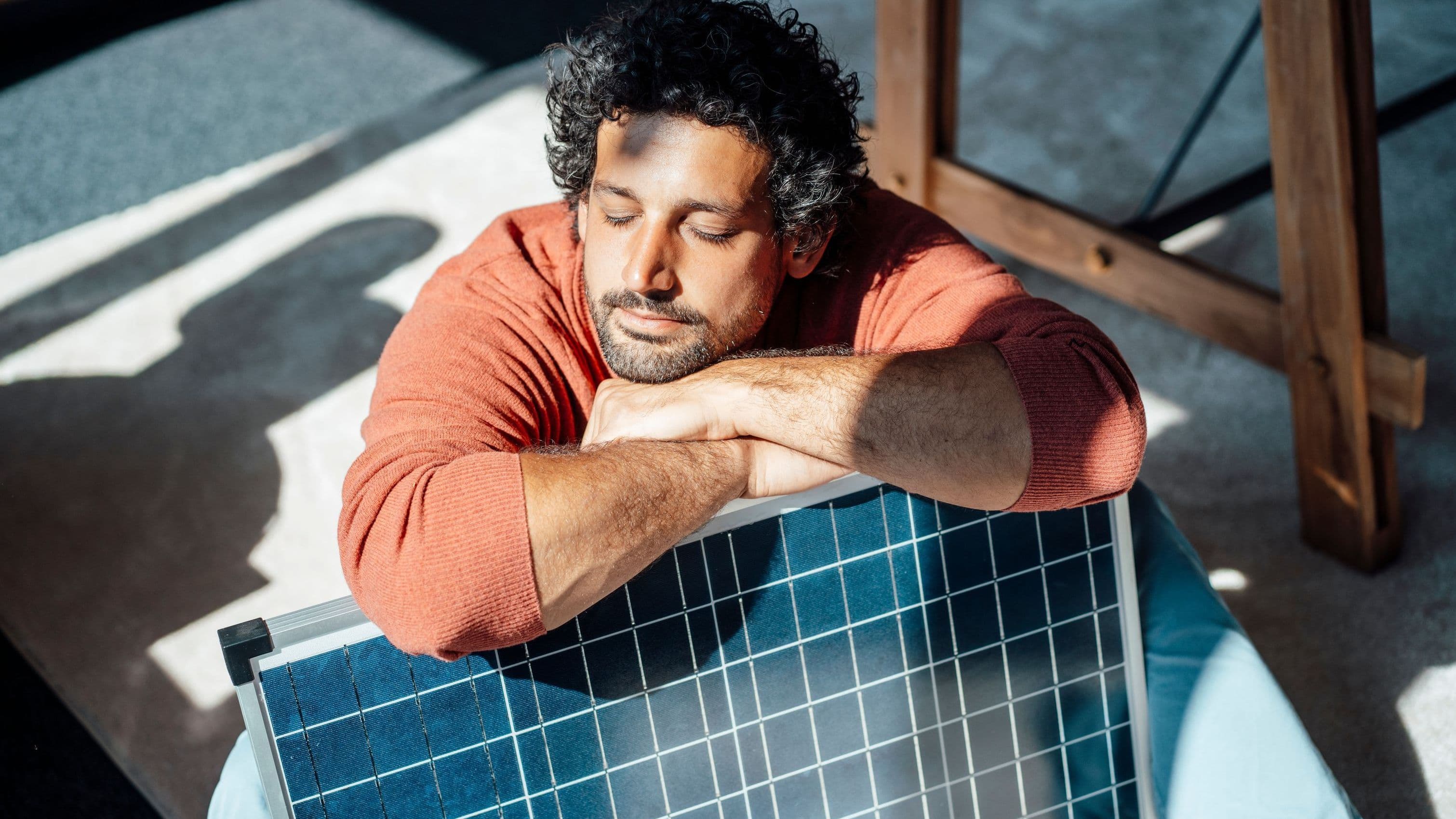 Ein Mann sitzt entspannt auf dem Boden und lehnt mit geschlossenen Augen an einem Solarmodul. Er trägt einen roten Pullover und genießt die Sonnenstrahlen, die durch das Fenster fallen.