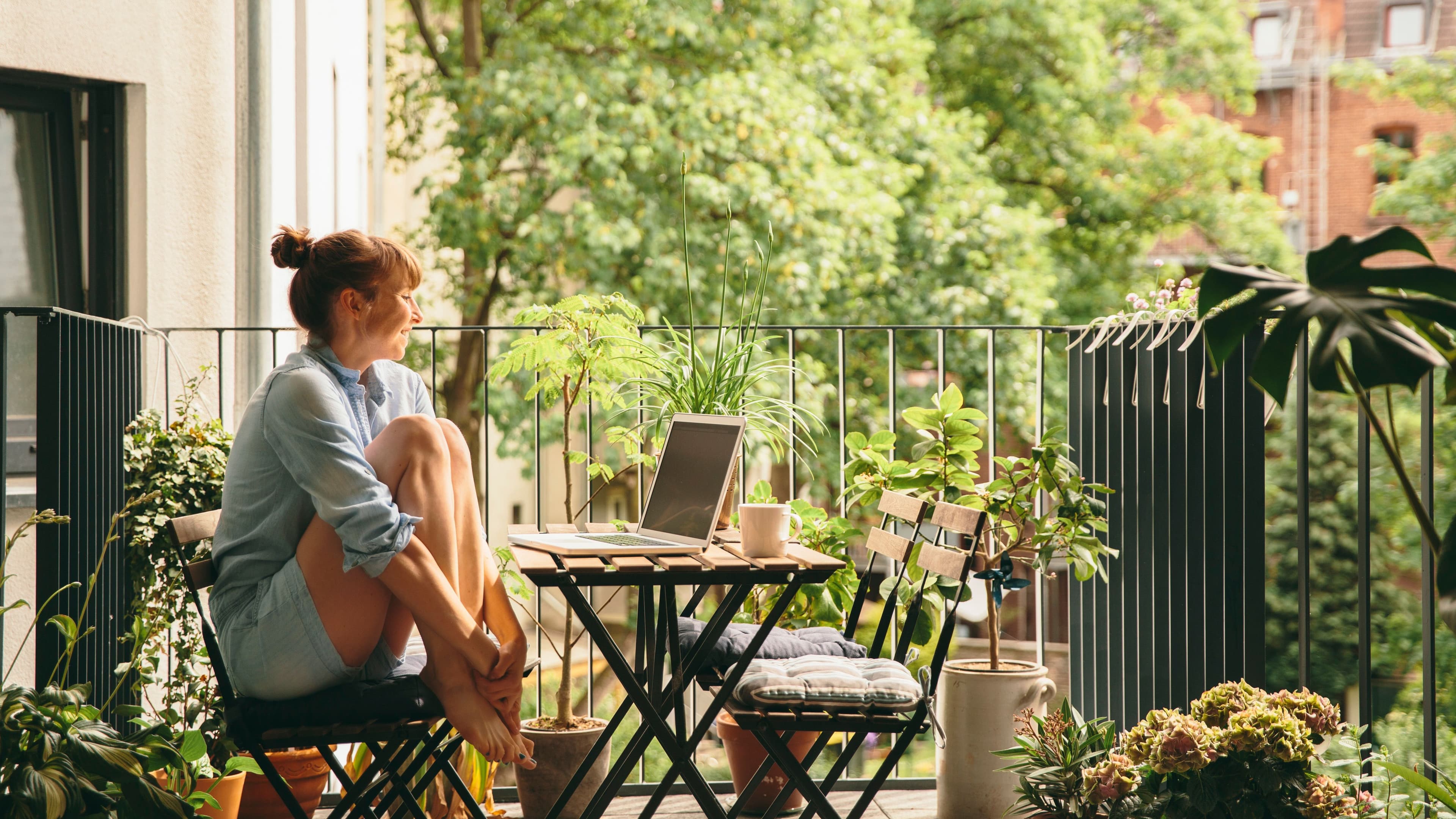 Frau sitzt entspannt auf einem Balkon an einem kleinen Tisch mit Laptop, umgeben von Pflanzen und Bäumen, und genießt die ruhige Atmosphäre.