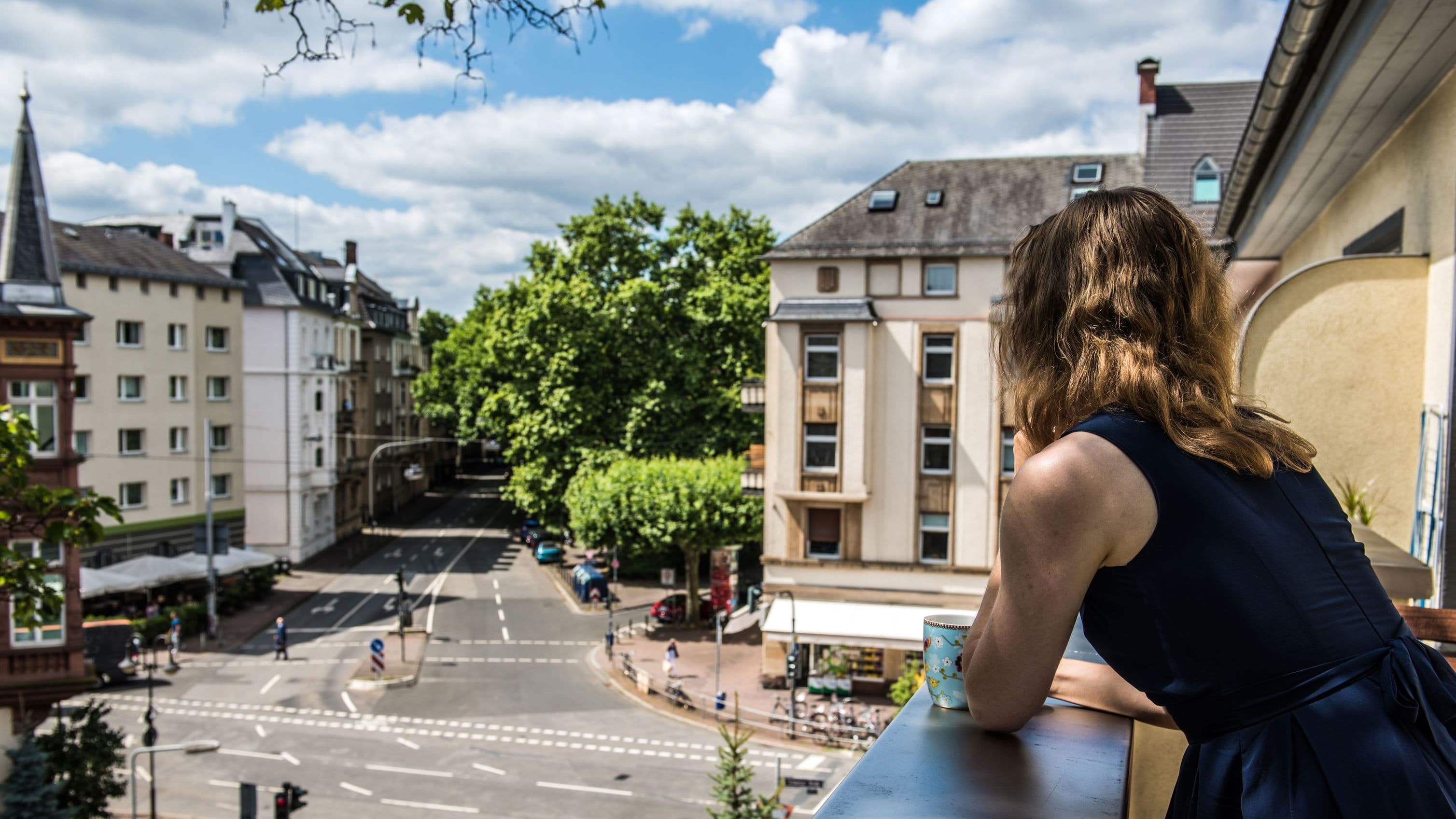 Eine Frau steht auf einem Balkon und blickt mit einer Tasse in der Hand auf eine ruhige Straßenszene mit Bäumen und Häusern in einer Stadt.