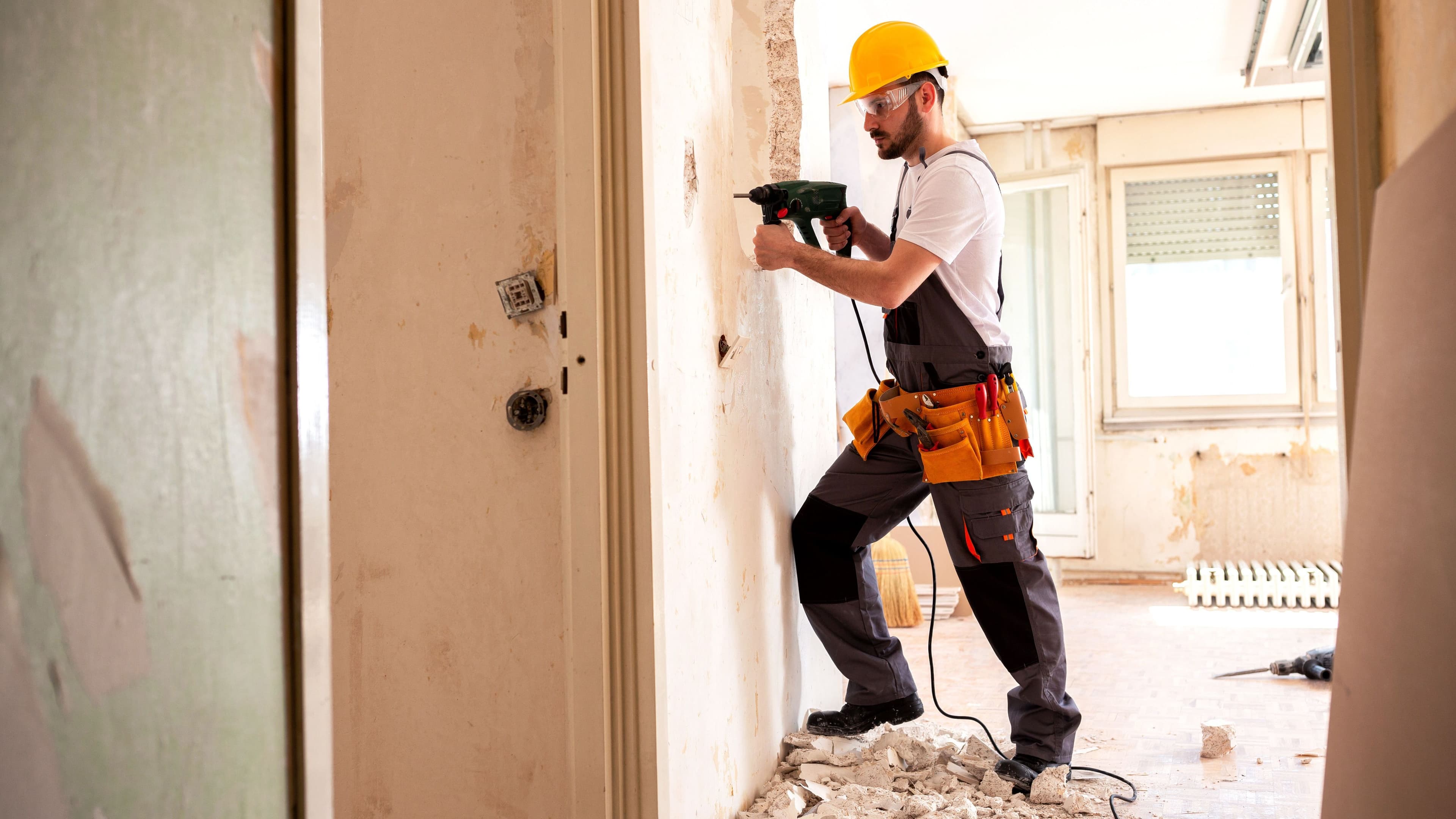 Handwerker mit Schutzhelm und Bohrmaschine arbeitet an einer Wand in einer Wohnung während einer Kernsanierung.