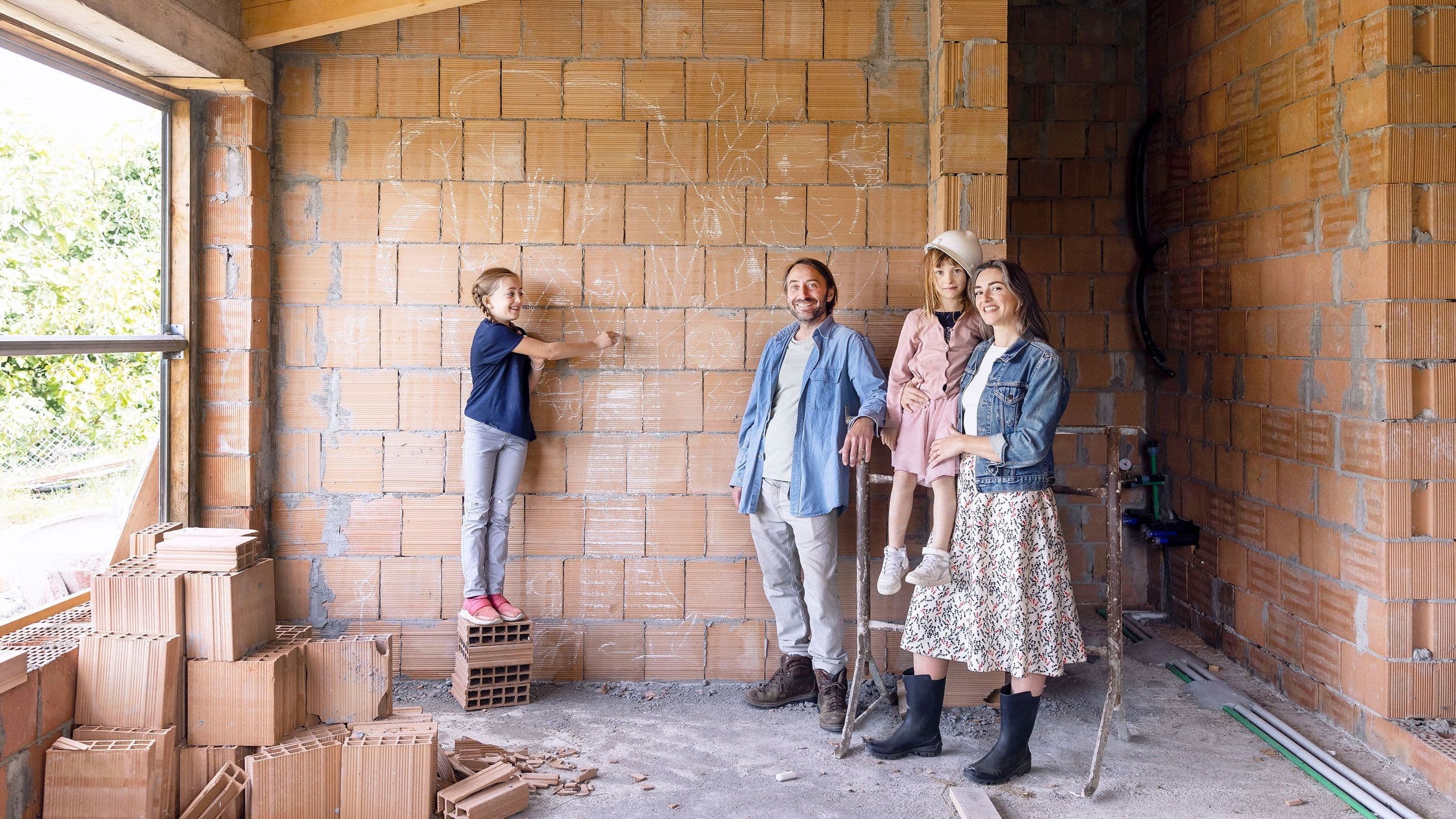 Eine vierköpfige Familie steht in dem Rohbau ihres neuen Hauses vor einer unverputzten Mauersteinwand und schaut in die Kamera. 