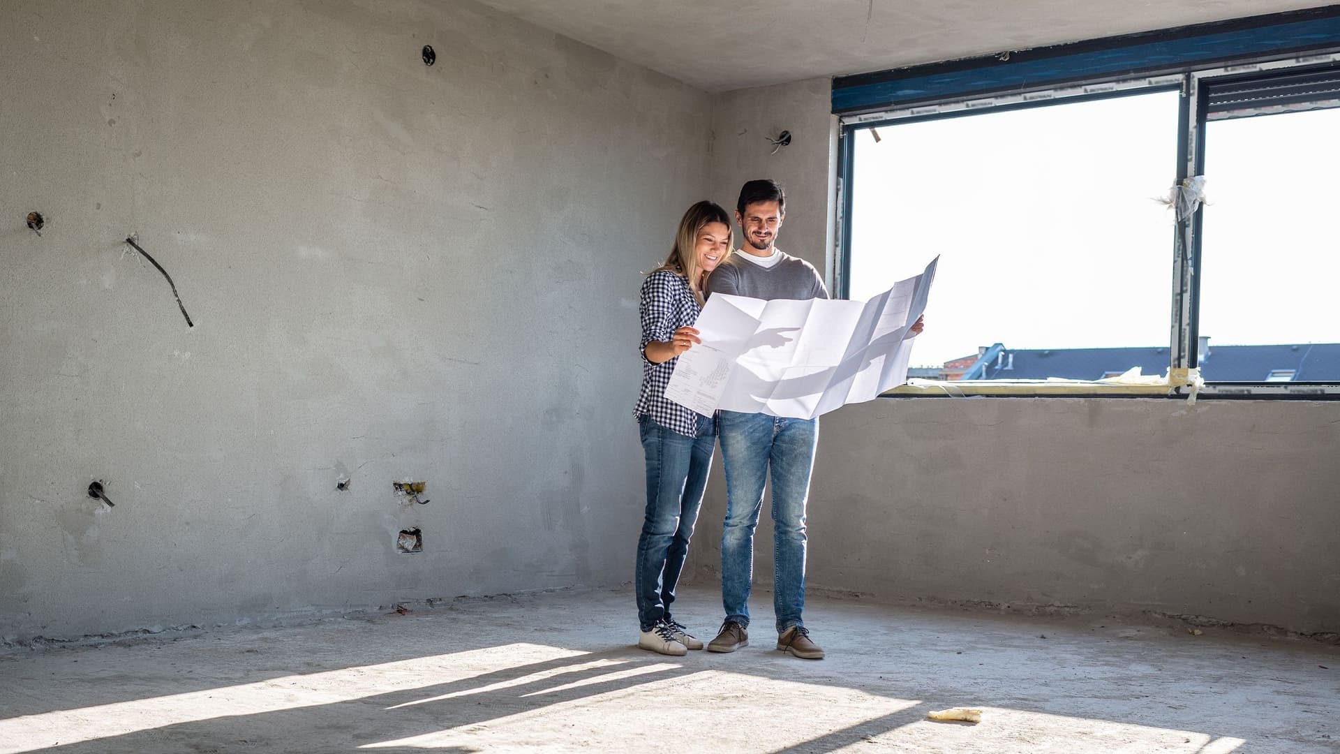 Eine Frau und ein Mann stehen inmitten eines Rohbaus auf einer Baustelle und sehen sich zusammen den Bauplan an. 