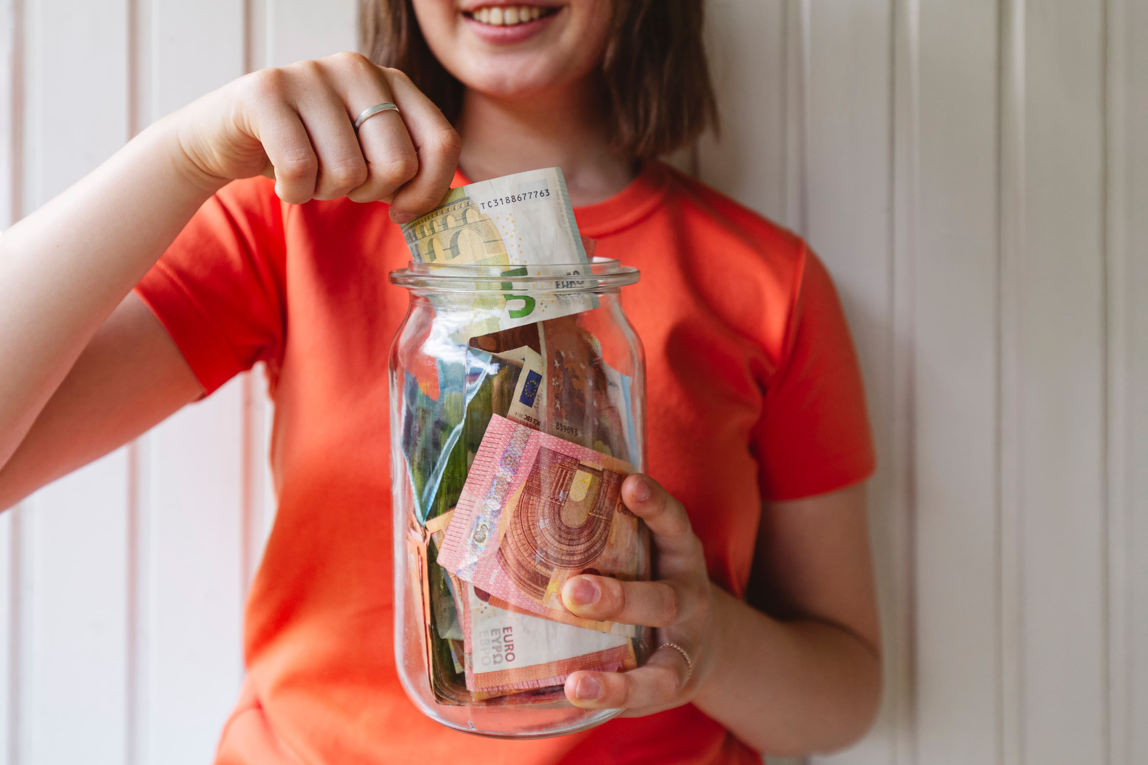 Oberkörper einer jungen Frau mit einem Glas voller Geld vor sich. mit einer Hand steckt sie Geld ins Glas.