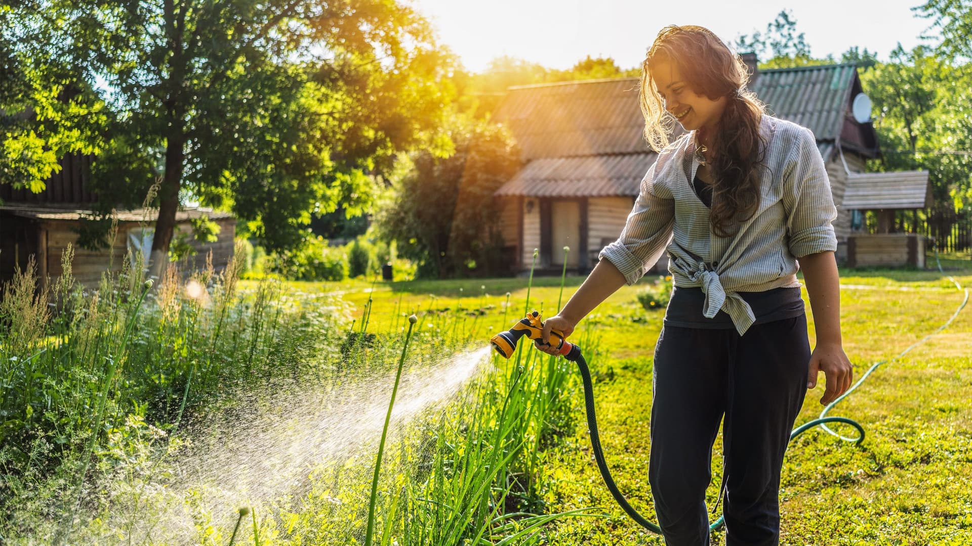Junge Frau bewässert mit einem Gartenschlauch Pflanzen im Garten. Im Hintergrund steht ein Landhaus.