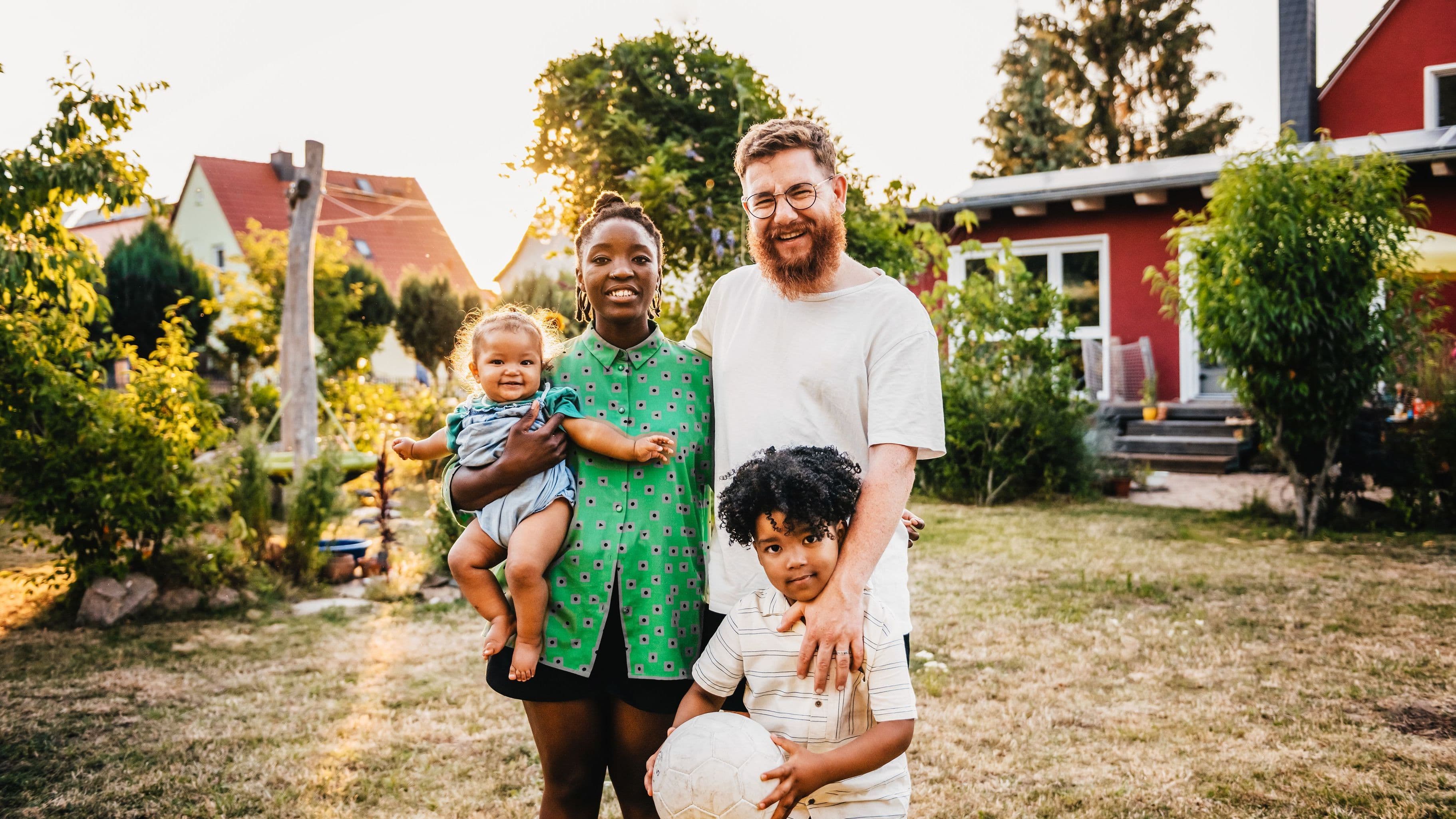 Eine Familie mit einem kleinen Kind und einem Baby steht vor einem roten Einfamilienhaus.