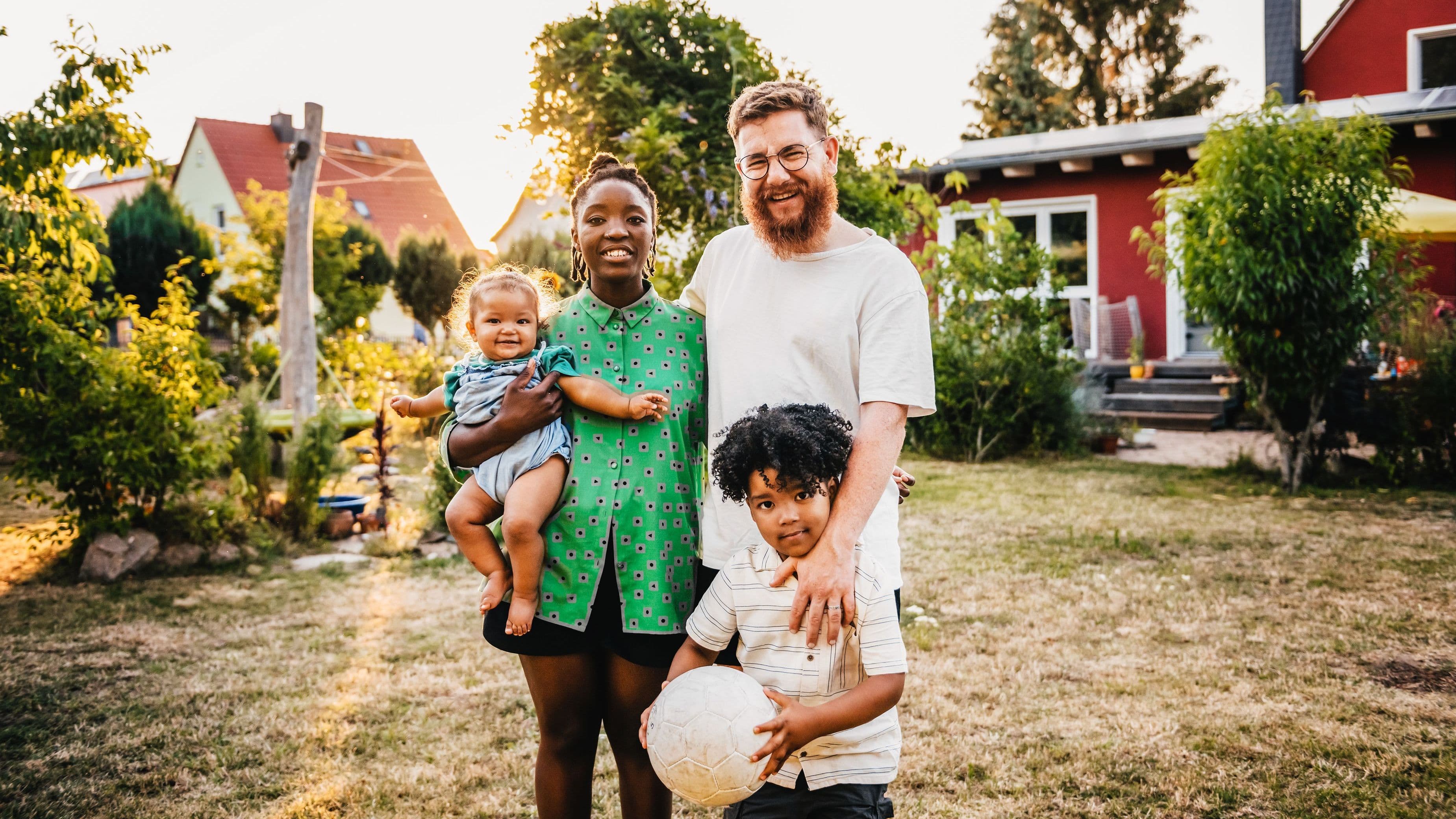 Eine Familie mit einem kleinen Kind und einem Baby steht vor einem roten Einfamilienhaus.