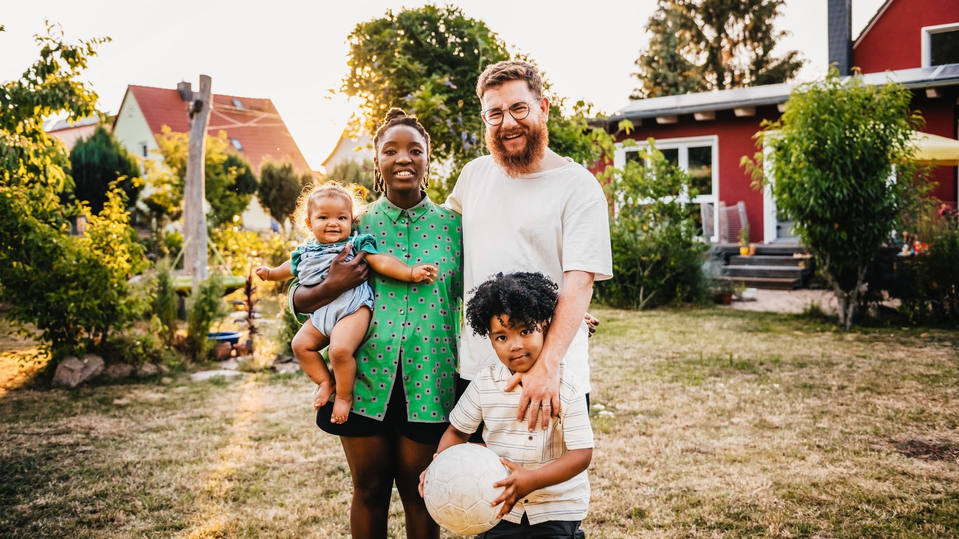 Eine Familie mit einem kleinen Kind und einem Baby steht vor einem roten Einfamilienhaus.