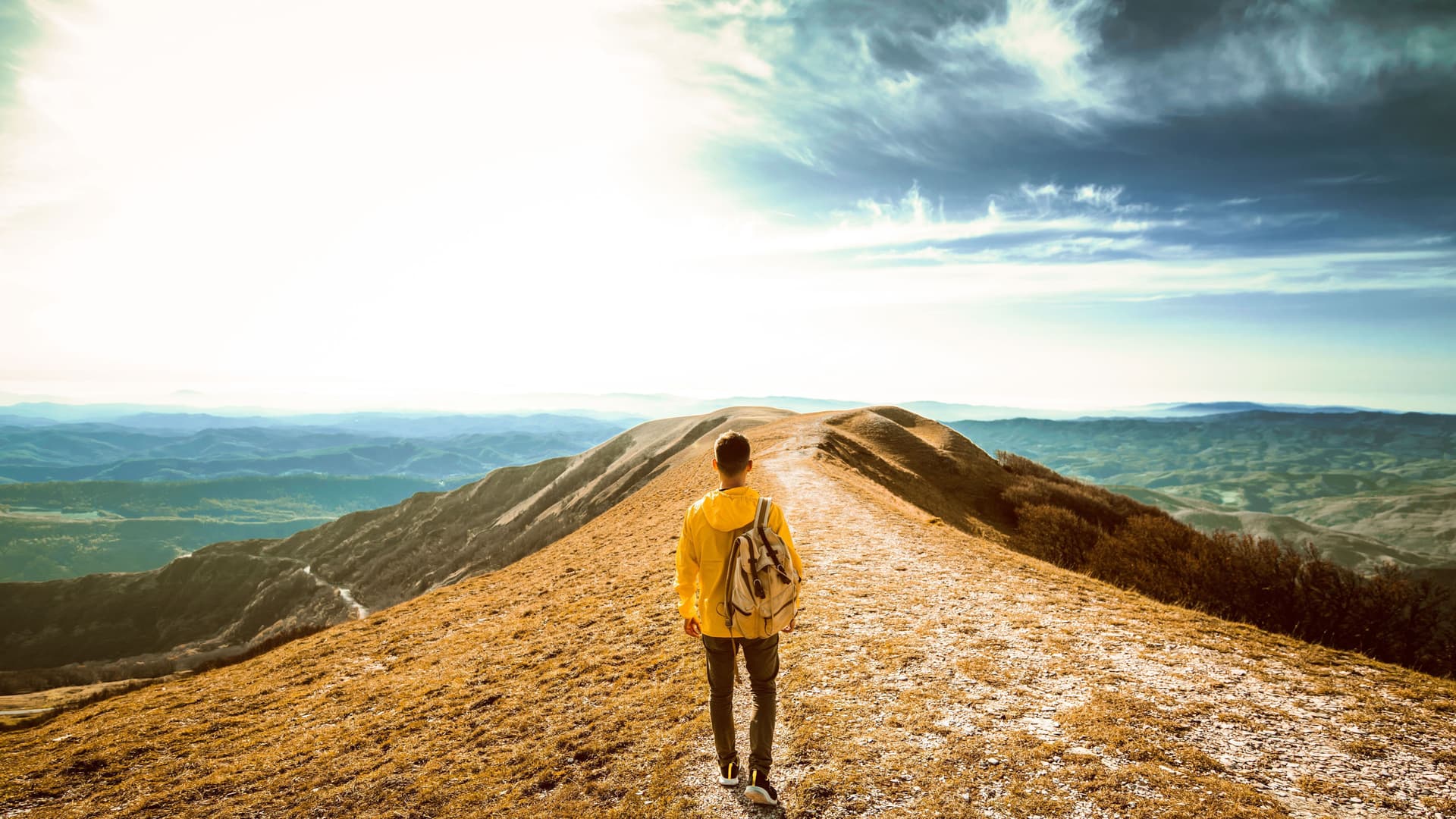 Wanderer läuft Richtung Bergspitze. Im Hintergrund dramatische Wolkenformationen und eine grüne Landschaft.