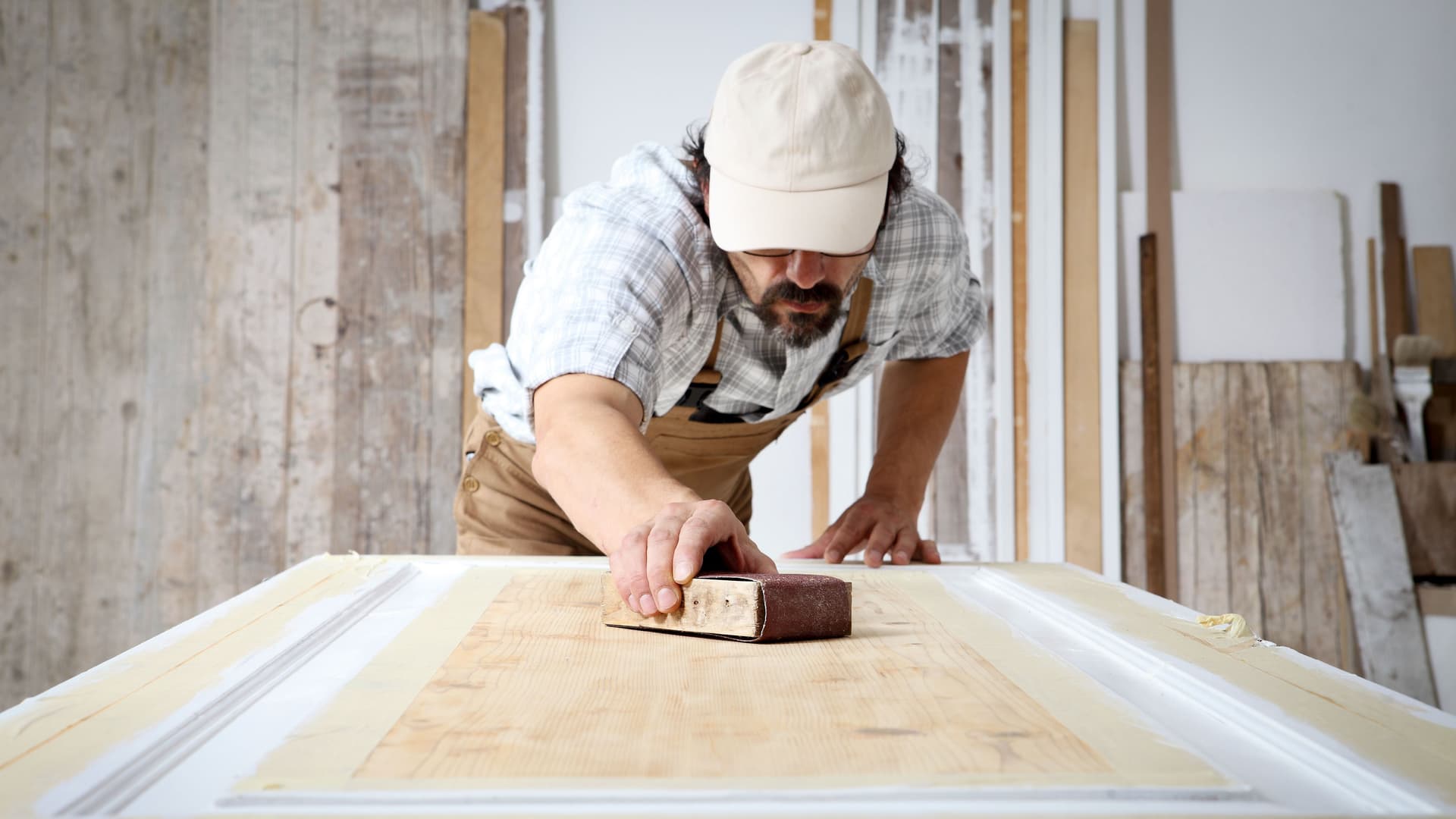 Schreiner mit Overall und Mütze, der in einer Schreinerei Holz bearbeitet und eine Holztür mit Schleifpapier abschleift.