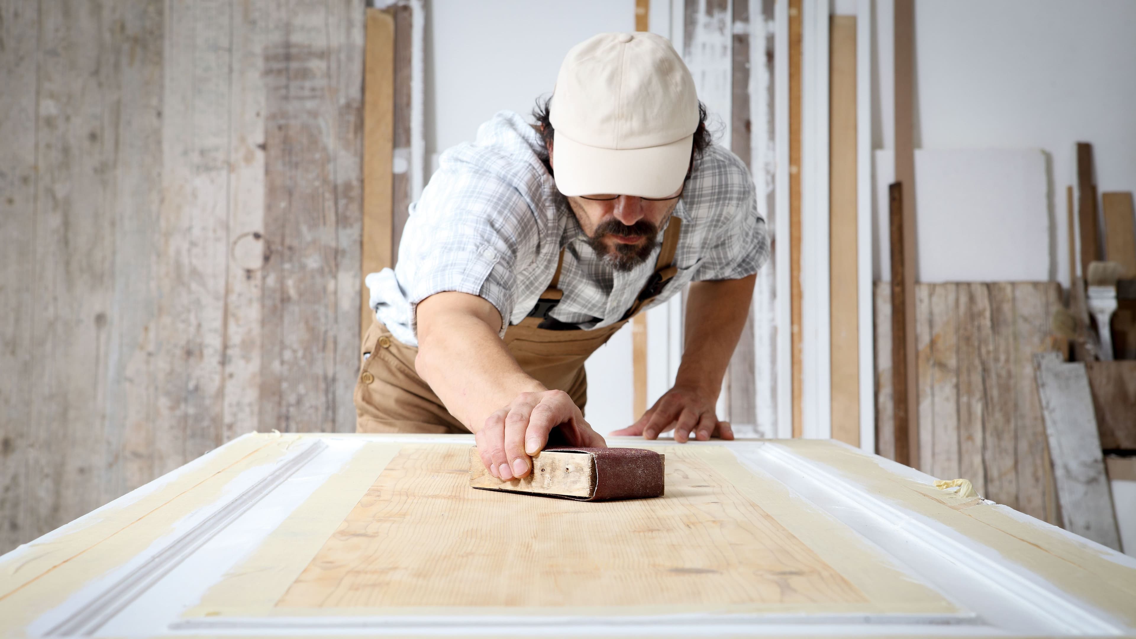Schreiner mit Overall und Mütze, der in einer Schreinerei Holz bearbeitet und eine Holztür mit Schleifpapier abschleift.
