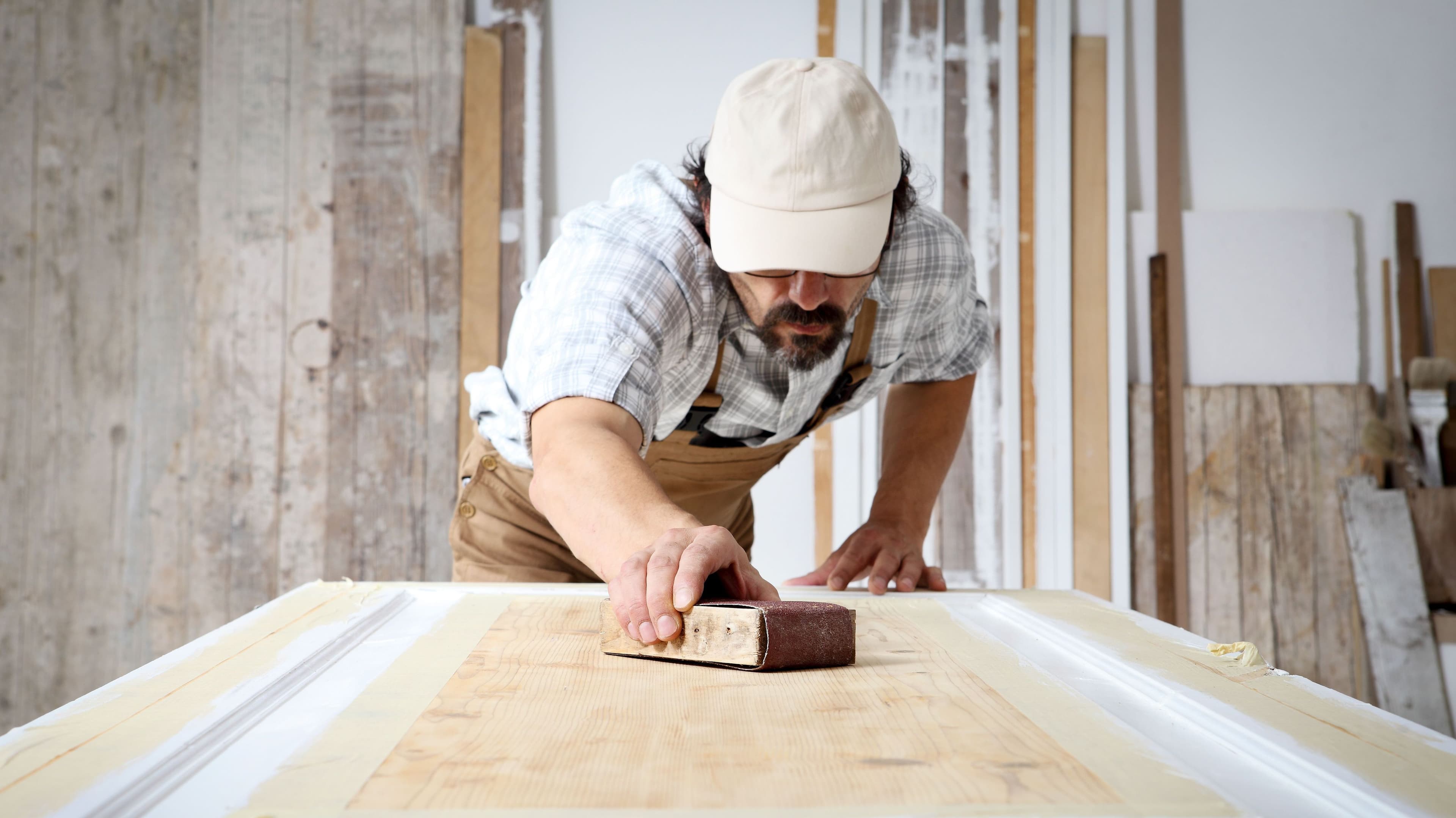 Schreiner mit Overall und Mütze, der in einer Schreinerei Holz bearbeitet und eine Holztür mit Schleifpapier abschleift.