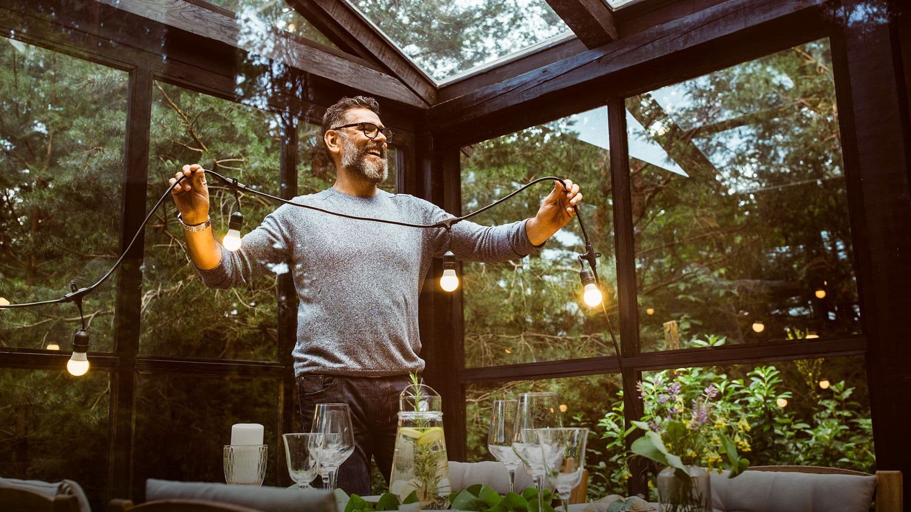 Ein Mann hängt in einem verglasten Wintergarten eine Lichterkette auf, während ein Tisch festlich gedeckt ist.