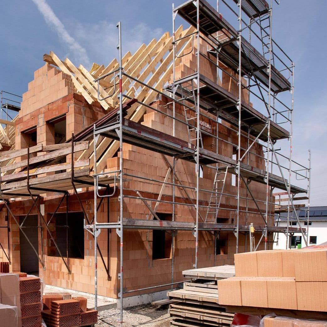 Ein im Bau befindliches Haus aus roten Ziegeln mit Gerüsten an der Fassade. Vor dem Gebäude liegen Stapel von Mauersteinen.