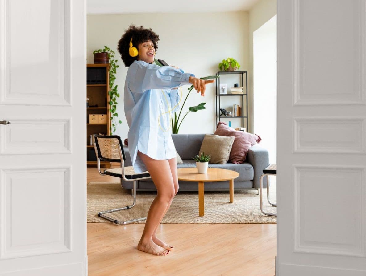 Glückliche Frau tanzt und singt ausgelassen mit Kopfhörern in ihrem Wohnzimmer.