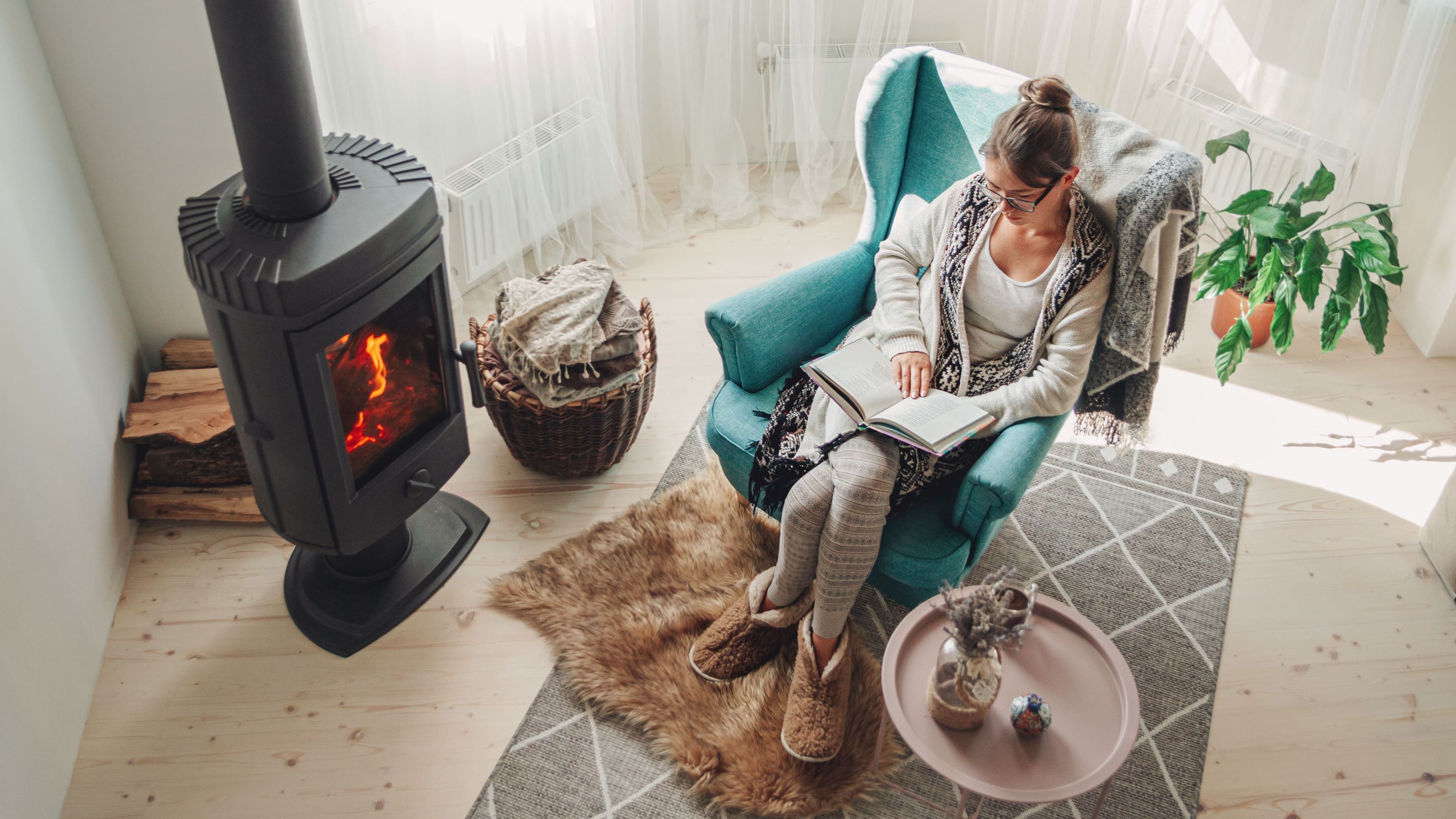 Blick von oben auf eine Frau im Wohnzimmer, die in einem Sessel vor dem Ofen sitzt und ein Buch liest.