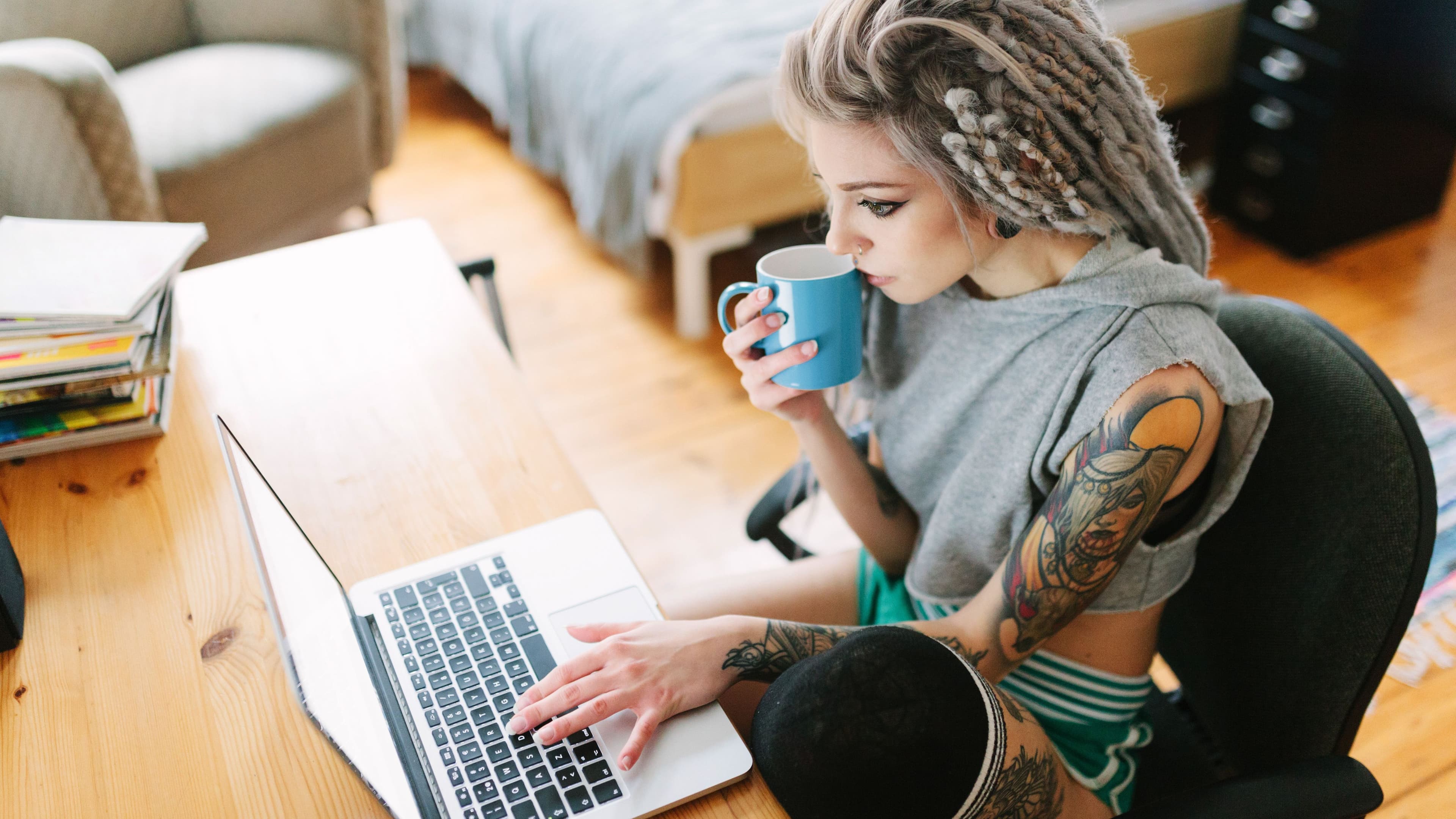 Eine junge tätowierte Frau mit grauen Dreadlocks sitzt konzentriert mit einer Tasse Kaffee vor ihrem Laptop.