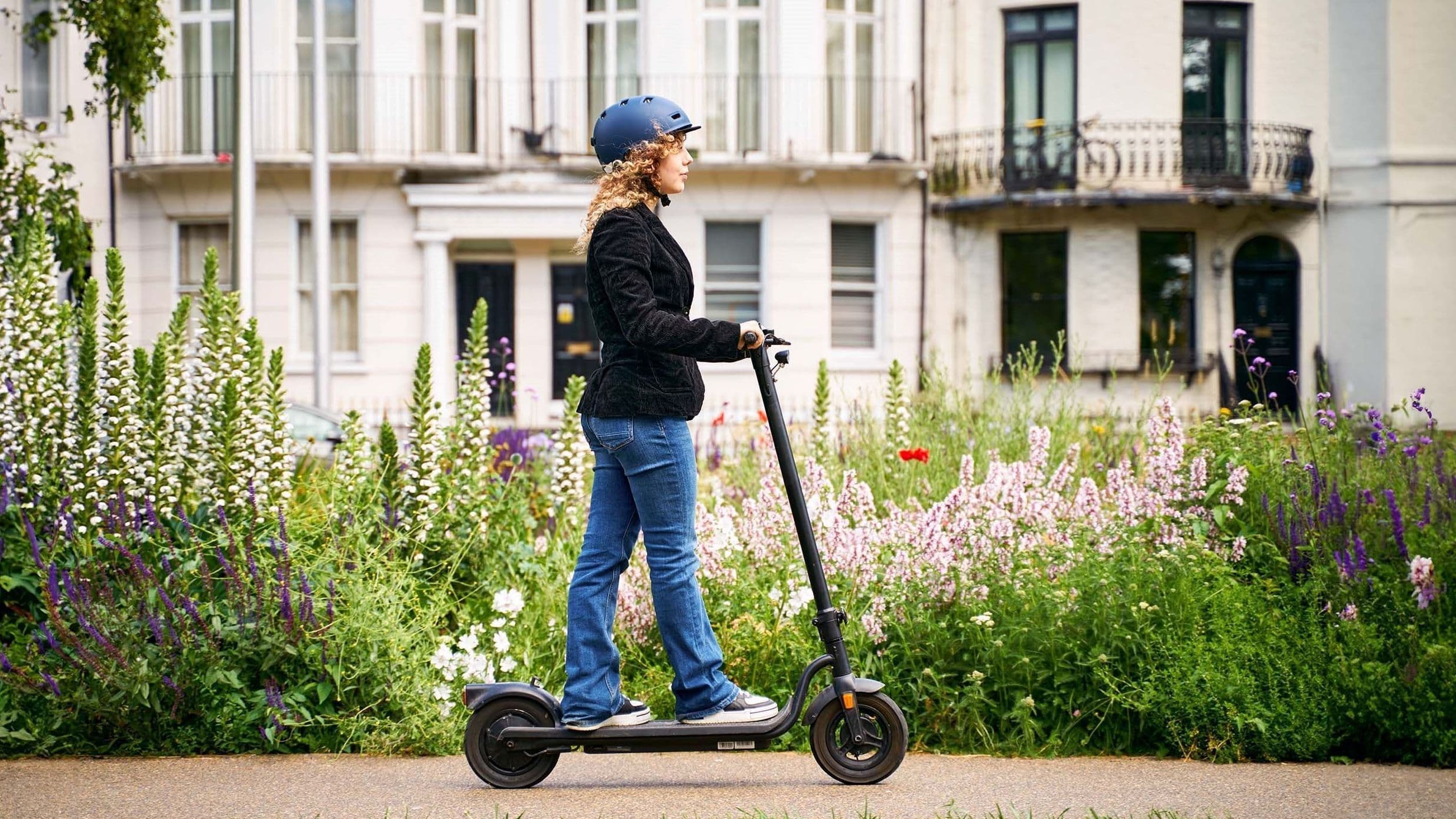 Eine junge Frau mit blauem Helm fährt auf einem E-Scooter einen Gehweg entlang. Sie trägt Jeans, Sneakers und eine dunkle Jacke. Im Hintergrund blühen hohe weiße, rosa und violette Blumen vor Stadthäusern mit Balkonen und großen Fenstern.