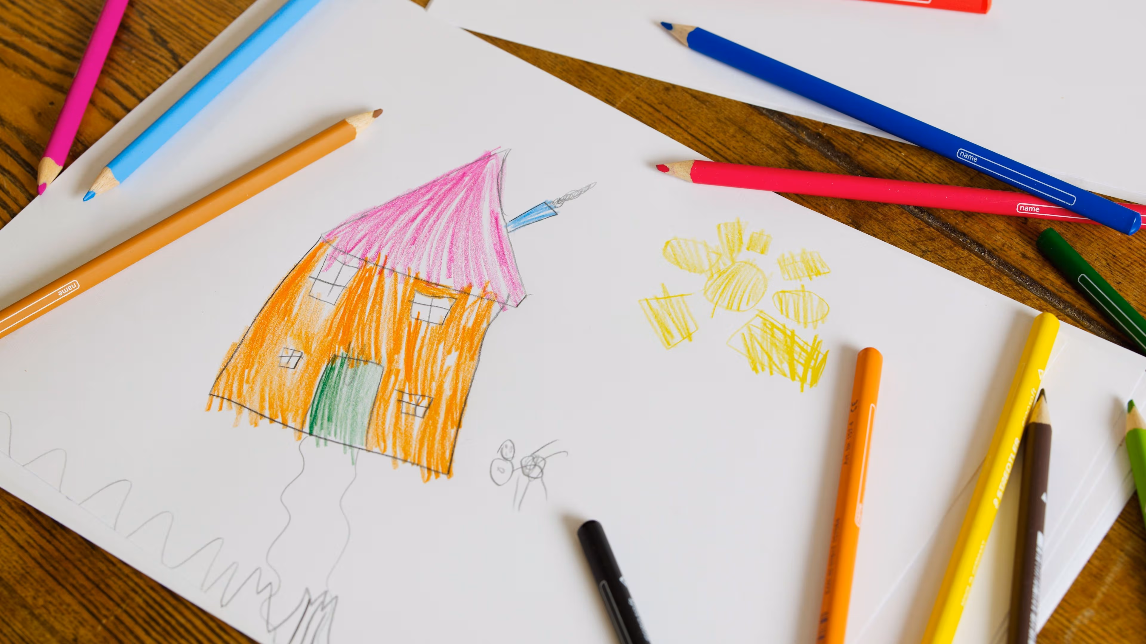 Ein Kinderzeichnung liegt mit Buntstiften auf einem Holztisch. Die Zeichnung zeigt ein Haus und eine Sonne.