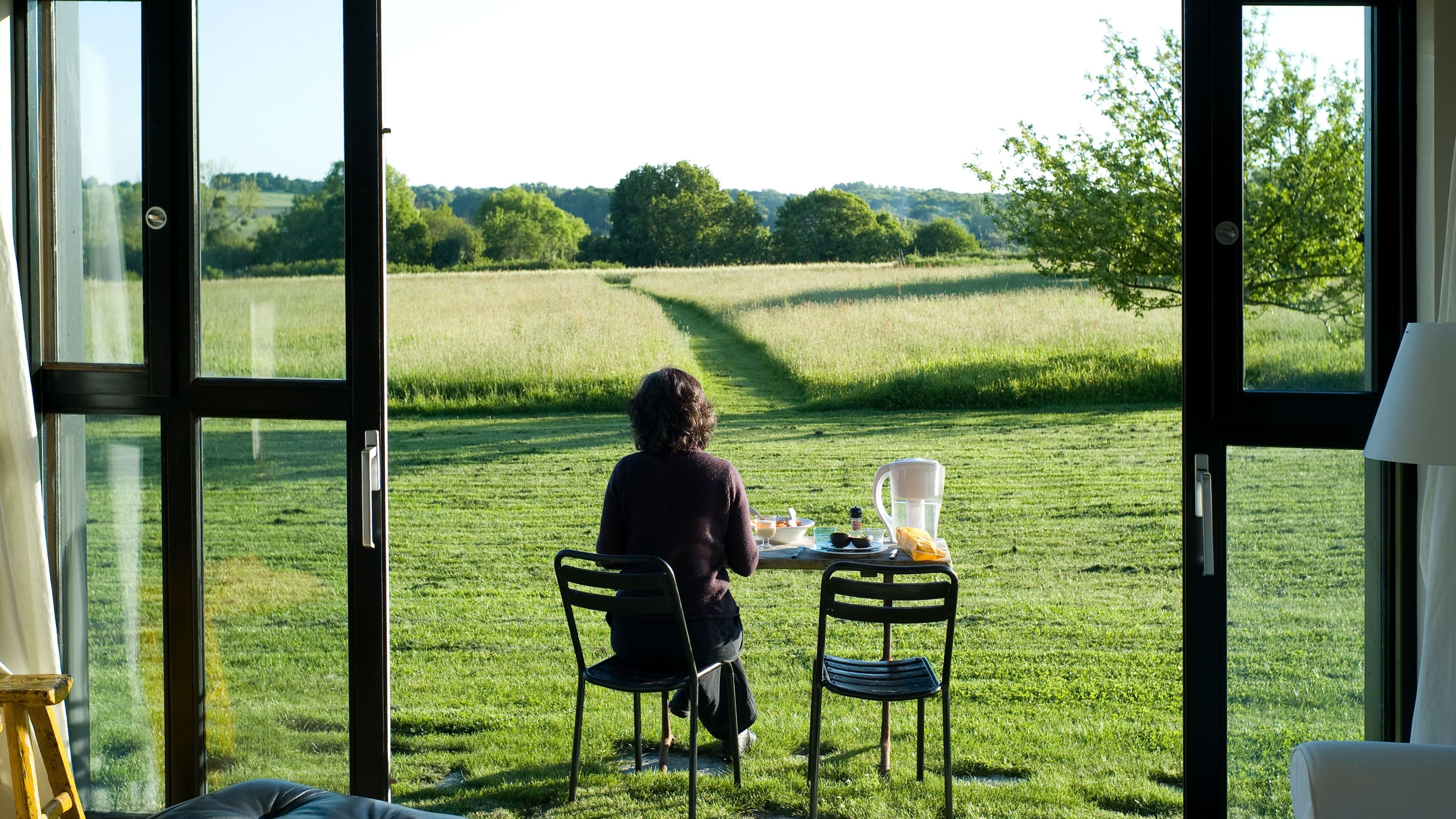 Ein Mann steht barfuß mit einer Tasse Kaffee und einem Smartphone am Fenster eines Tiny Houses und blickt dabei in die Natur.
