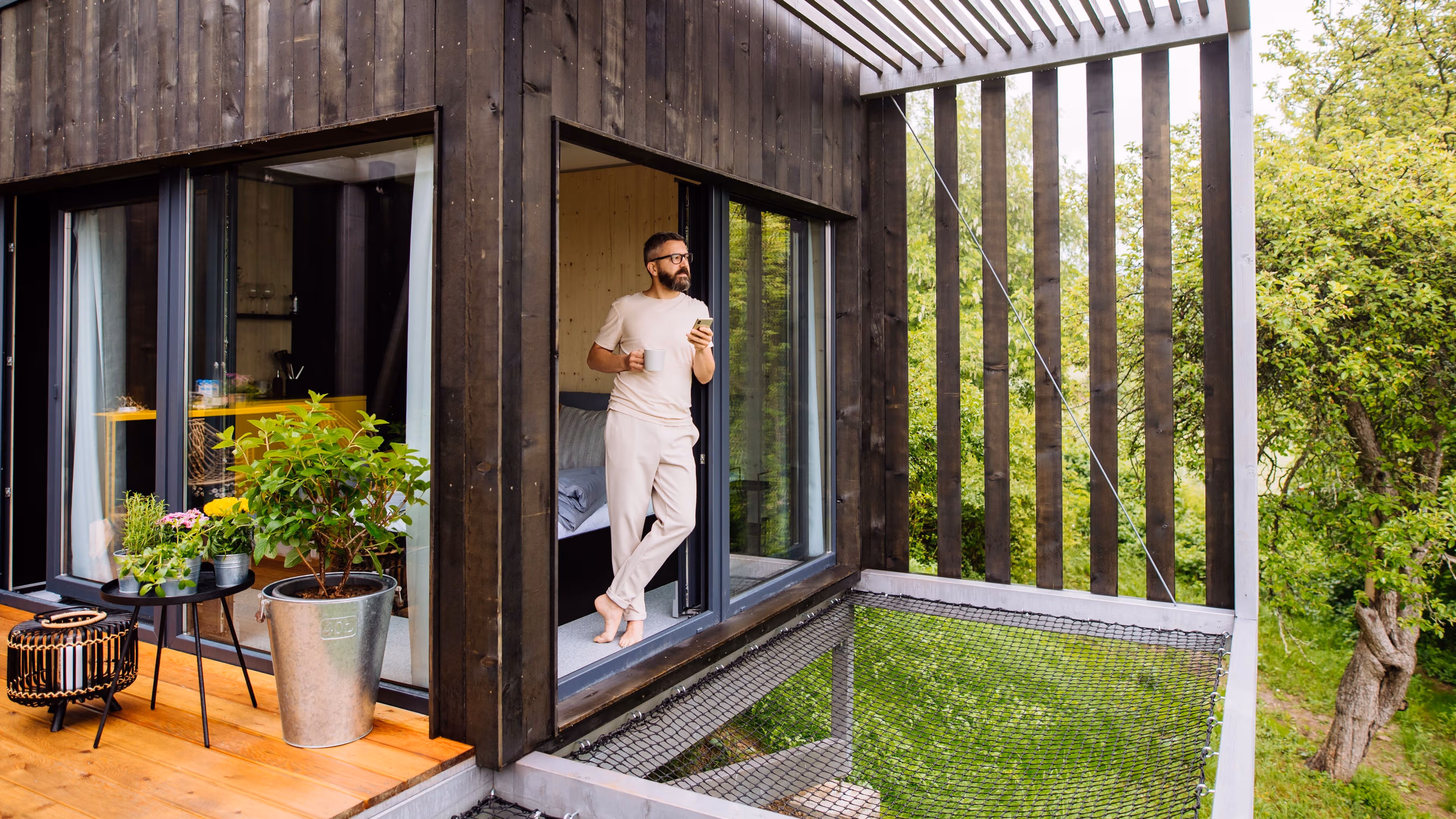 Ein Mann steht barfuß mit einer Tasse Kaffee und einem Smartphone am Fenster eines Tiny Houses und blickt dabei in die Natur.