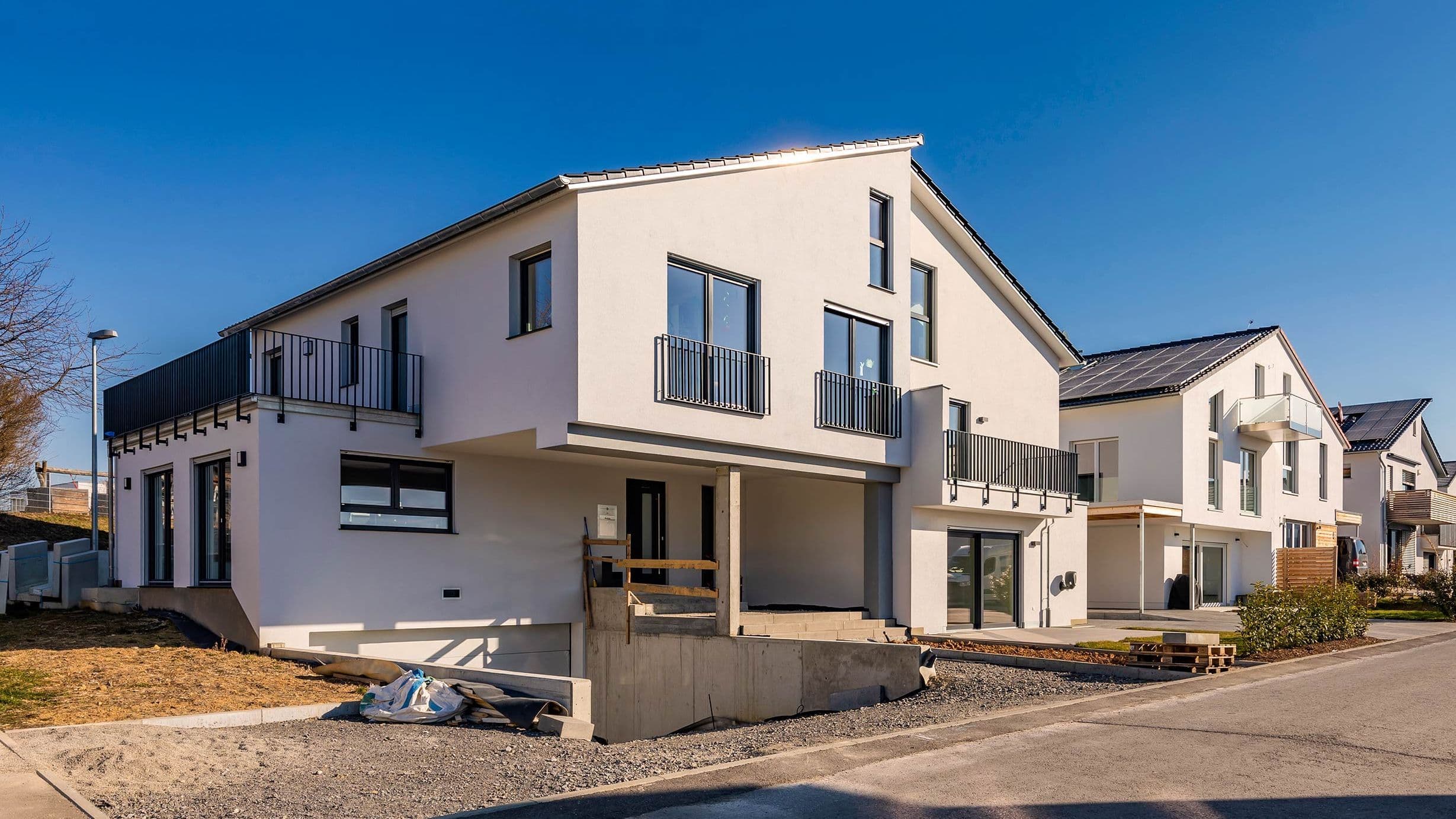 Eine Außenaufnahme eines Straßenzuges mit neugebauten Einfamilienhäusern. Ein weißes Haus ist zentral im Bild. Ähnliche Häuser reihen sich anhand einer Fluchtlinie hintereinander.
