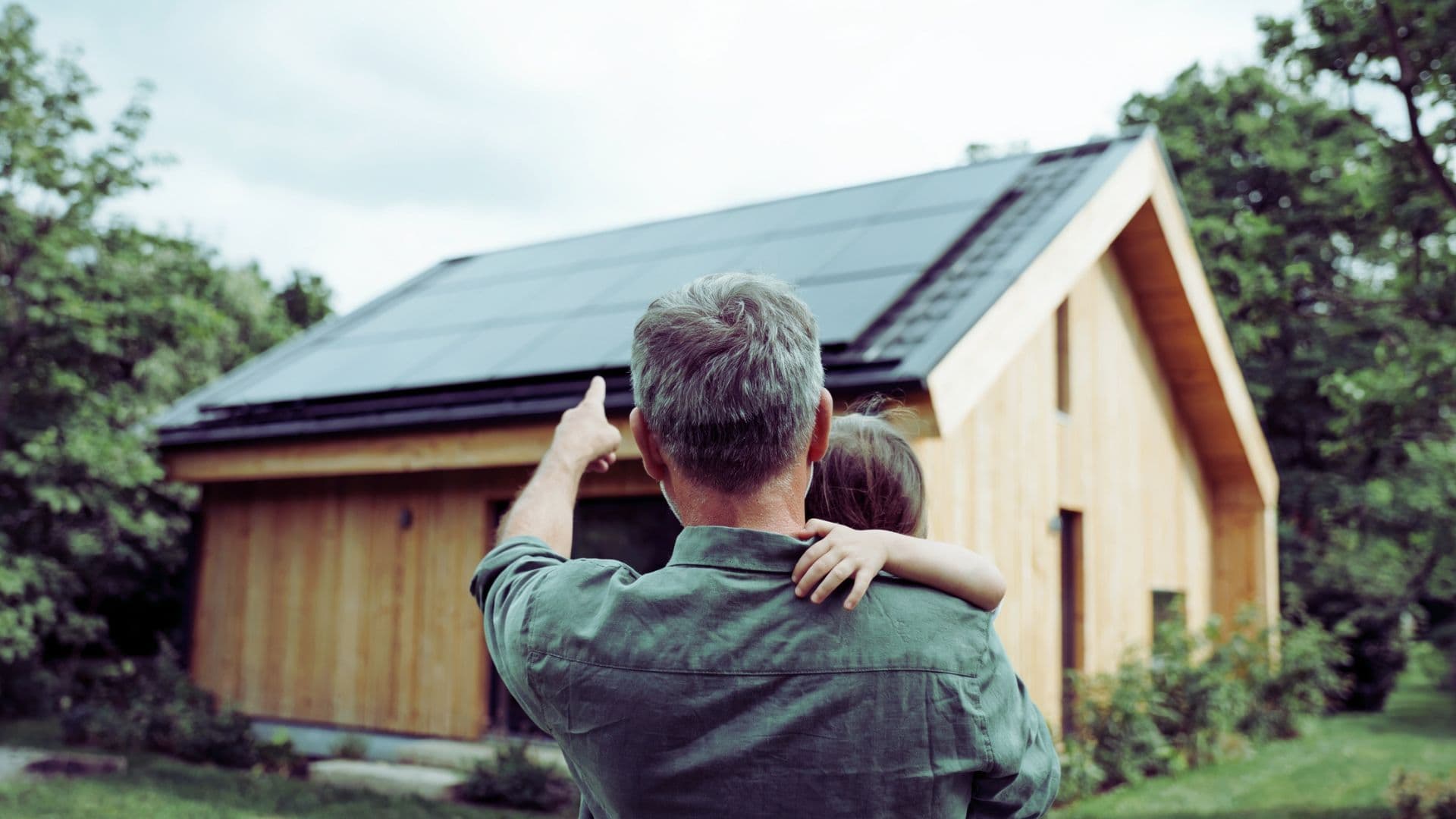 Vater trägt sein Kind auf den Armen und zeigt auf ein Haus mit Solaranlage auf dem Dach.