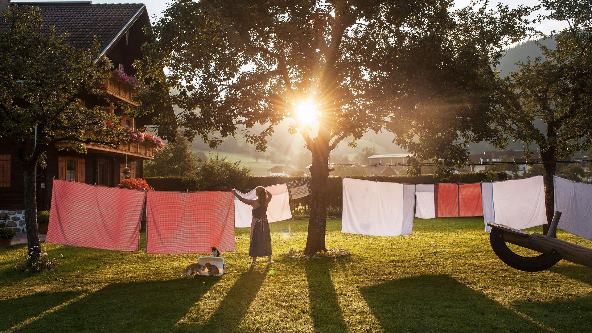 Ein Frau hängt in einem Garten Wäsche auf eine Wäscheleine. Im Hintergrund steht ein traditionelles österreichisches Haus. Die Sonne scheint und es ist warm.