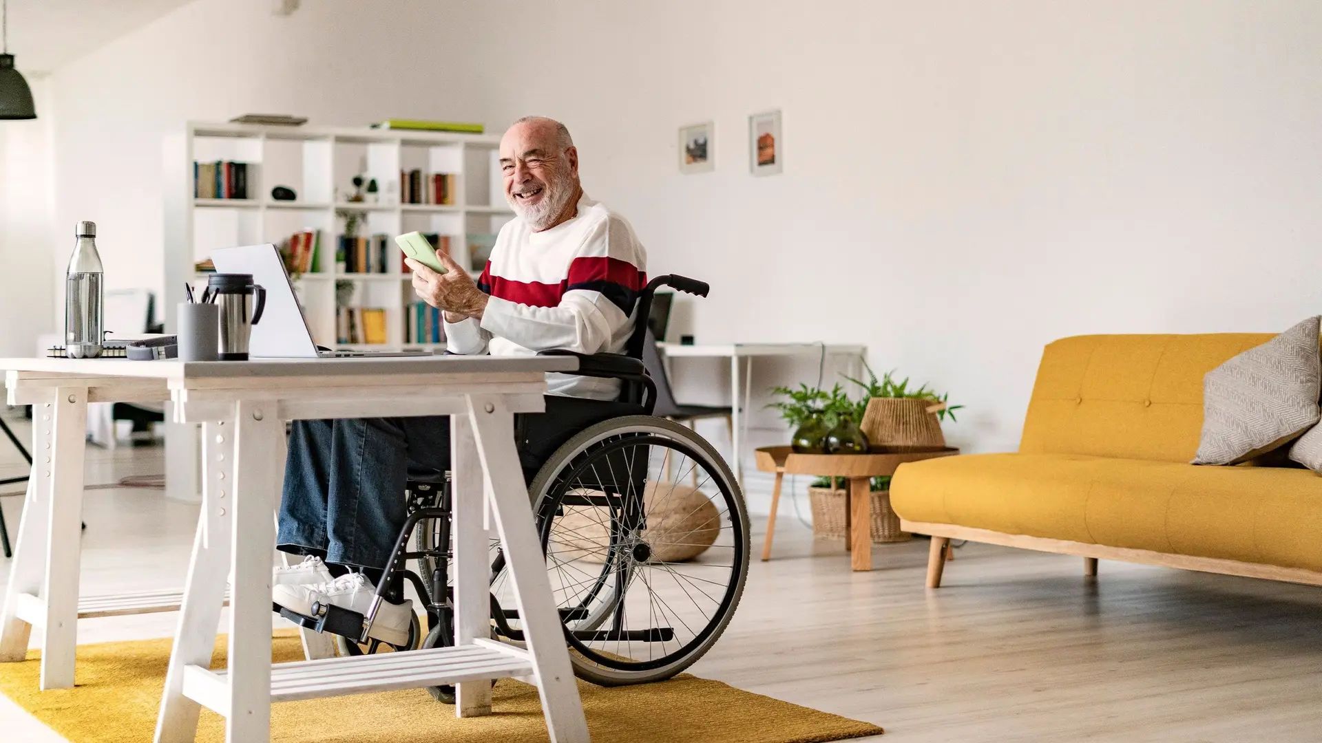 Älterer Mann im Rollstuhl sitzt mit seinem Smartphone an seinem Schreibtisch.