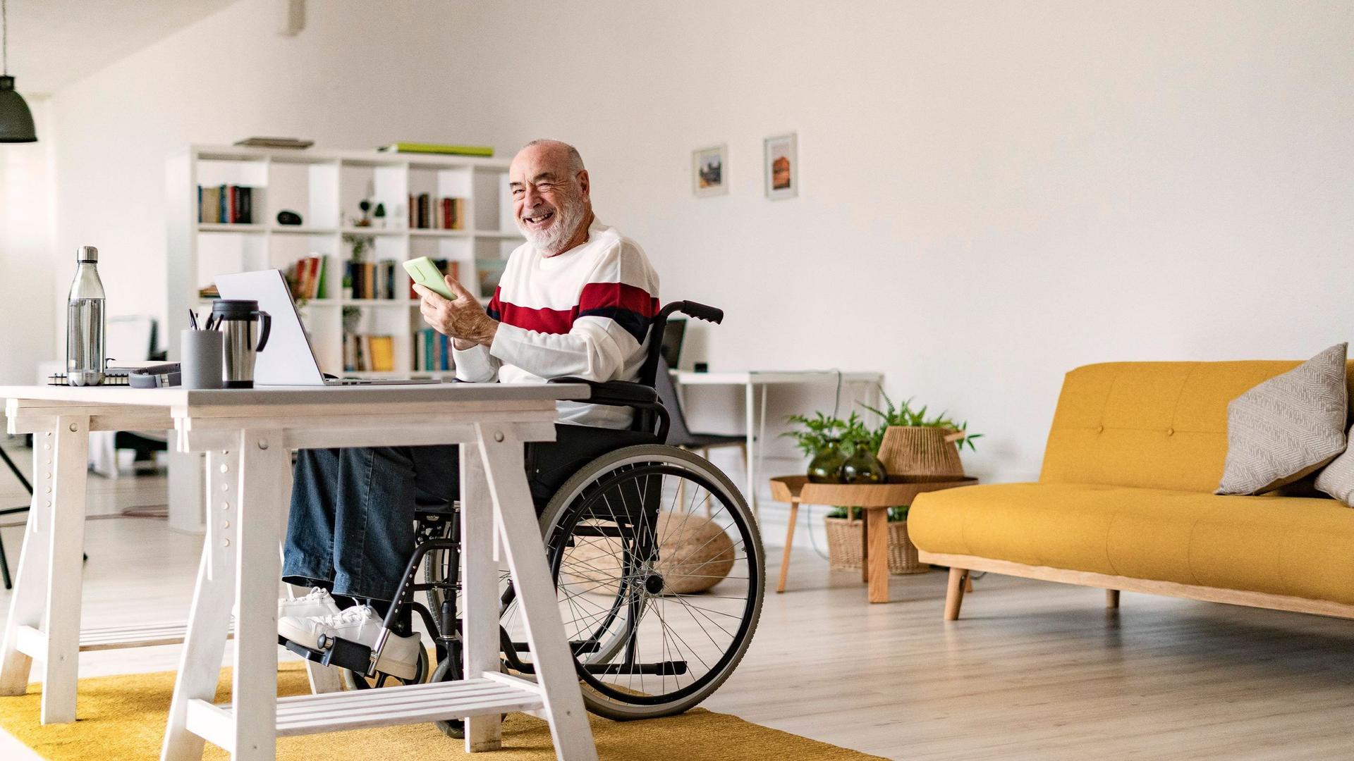 Älterer Mann im Rollstuhl sitzt mit seinem Smartphone an seinem Schreibtisch.