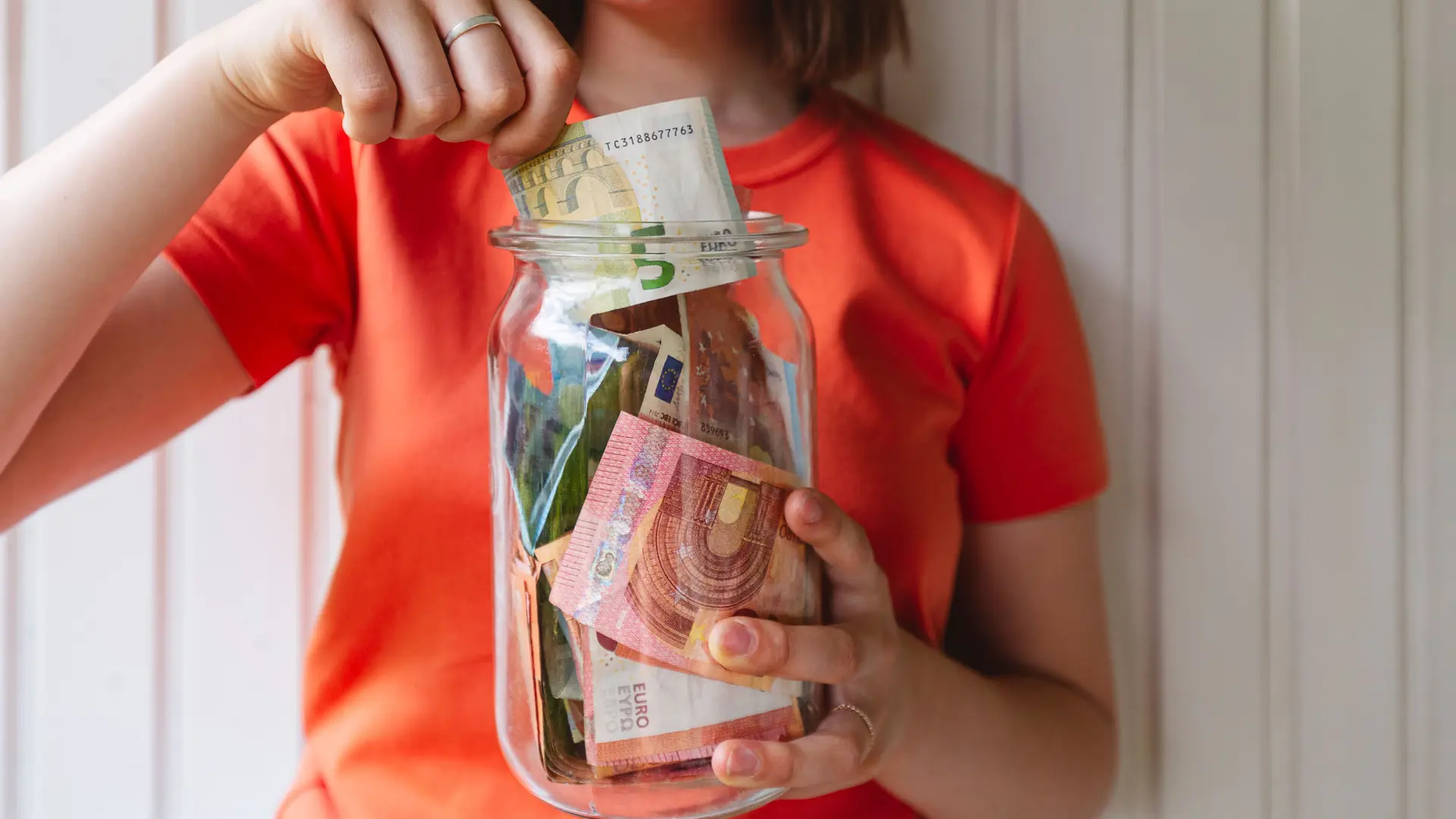 Oberkörper einer jungen Frau mit einem Glas voller Geld vor sich. mit einer Hand steckt sie Geld ins Glas.
