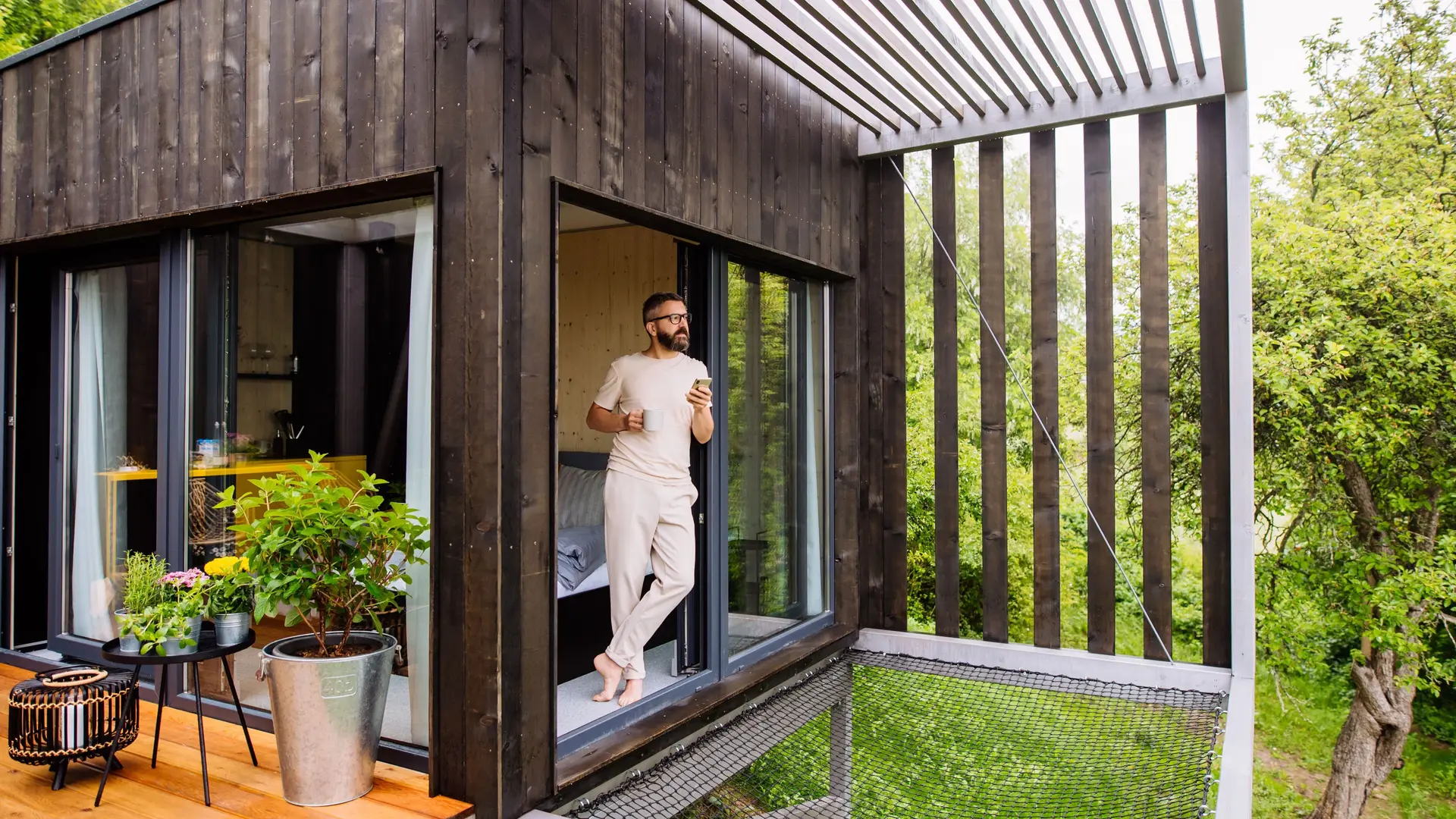 Ein Mann steht barfuß mit einer Tasse Kaffee und einem Smartphone am Fenster eines Tiny Houses und blickt dabei in die Natur.