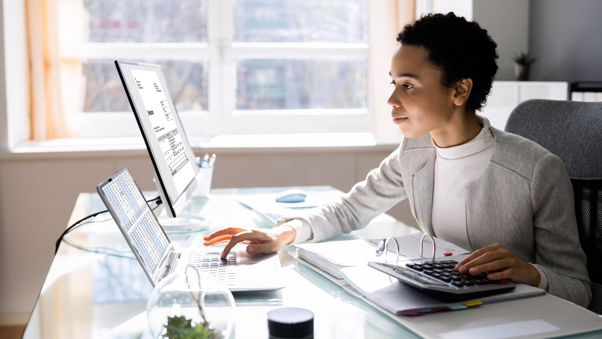Eine Frau sitzt mit einem Rechner vor einem Laptop und einem Bildschirm. Mit der linken Hand bedient sie den Rechner, mit der rechten Hand tippt sie am Laptop.